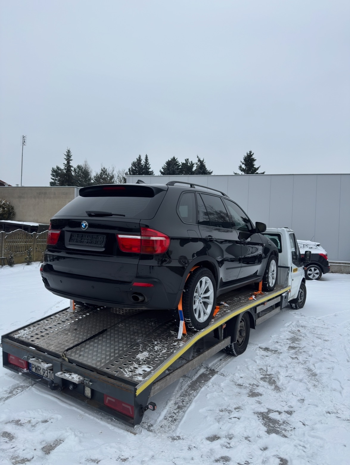 Czarny SUV BMW na lawecie w zimowej scenerii. Auto zabezpieczone pasami transportowymi, widoczny fragment drugiego pojazdu w tle. Śnieg na ziemi i pochmurne niebo.