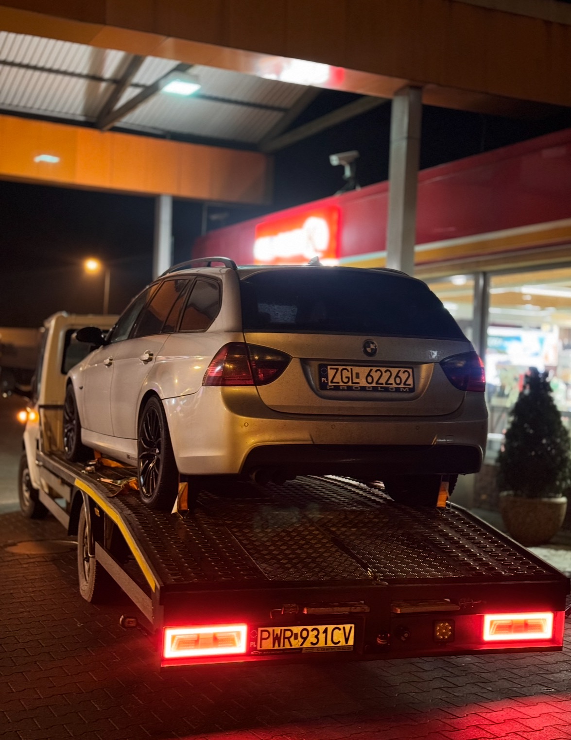 Srebrne BMW kombi na platformie lawety, widok z tyłu, nocne oświetlenie, tablice rejestracyjne widoczne. Auto z czarnymi felgami.