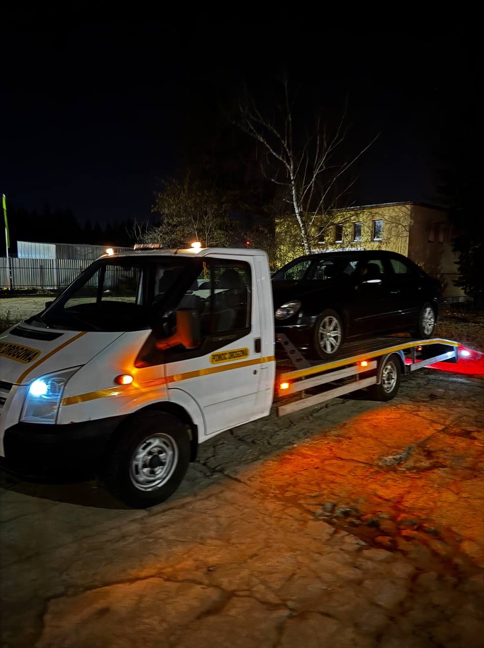 Nocny transport czarnego samochodu osobowego na lawecie pomocy drogowej. Biały bus z żółtymi pasami i napisem 'Pomoc Drogowa'.