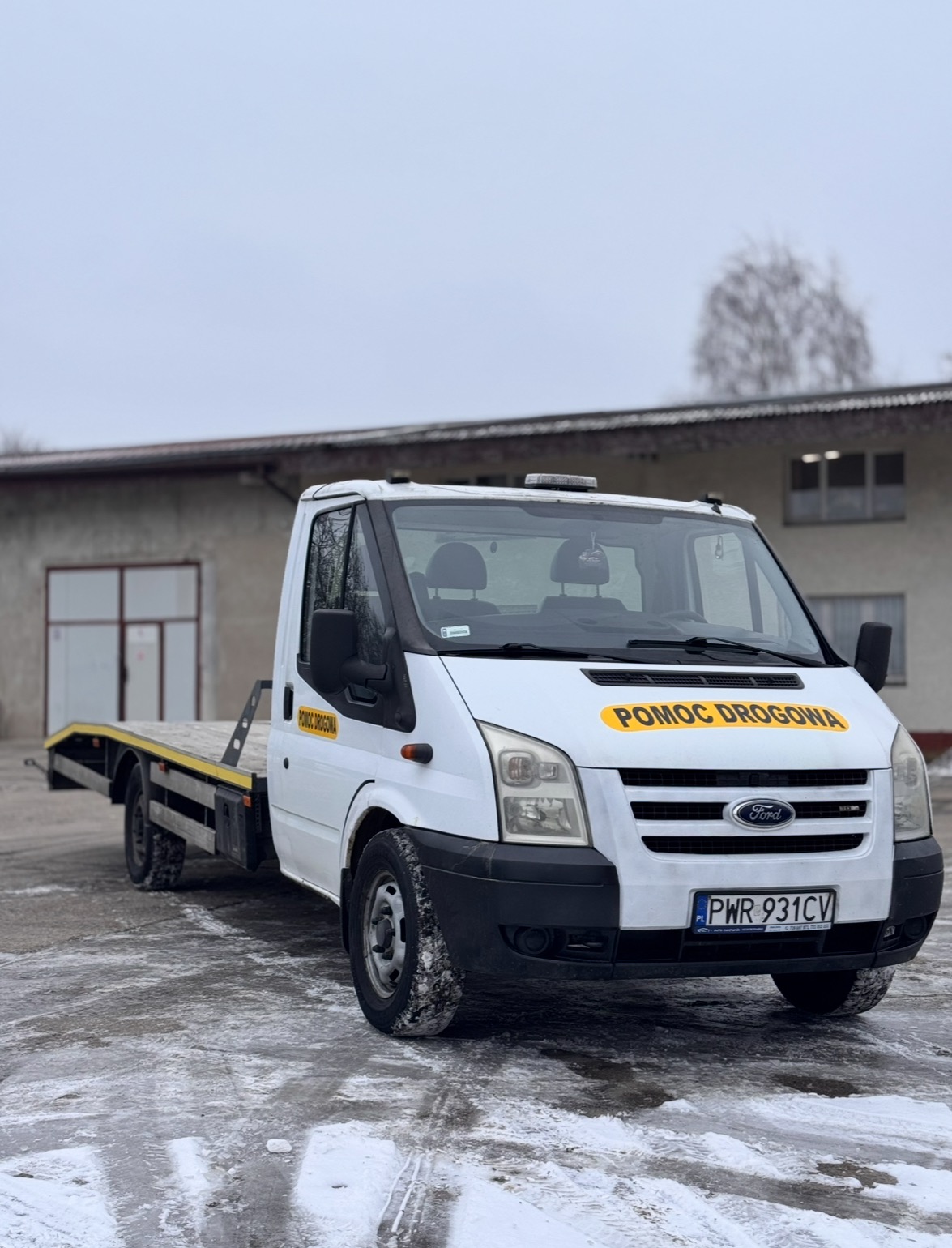 Biała laweta Ford Transit z żółtym napisem 'Pomoc Drogowa' na tle szarego budynku i zimowego krajobrazu. Widoczny numer rejestracyjny PWR 931CV.