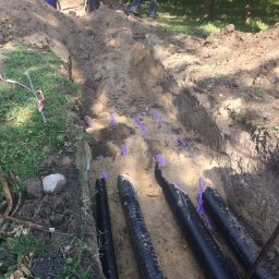 Zakład Budowlany - Stanisław Andrysiewicz - Wykop z ułożonymi czarnymi rurami, zabezpieczonymi fioletową taśmą. W tle widoczni pracownicy w trakcie prac ziemnych. Prace instalacyjne w terenie.