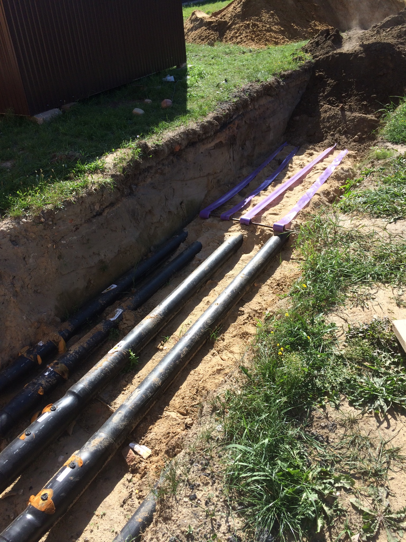 Rury w wykopie, zabezpieczone fioletowymi taśmami. Widoczne połączenia rur i ziemia z trawą. Instalacja podziemna w trakcie realizacji.