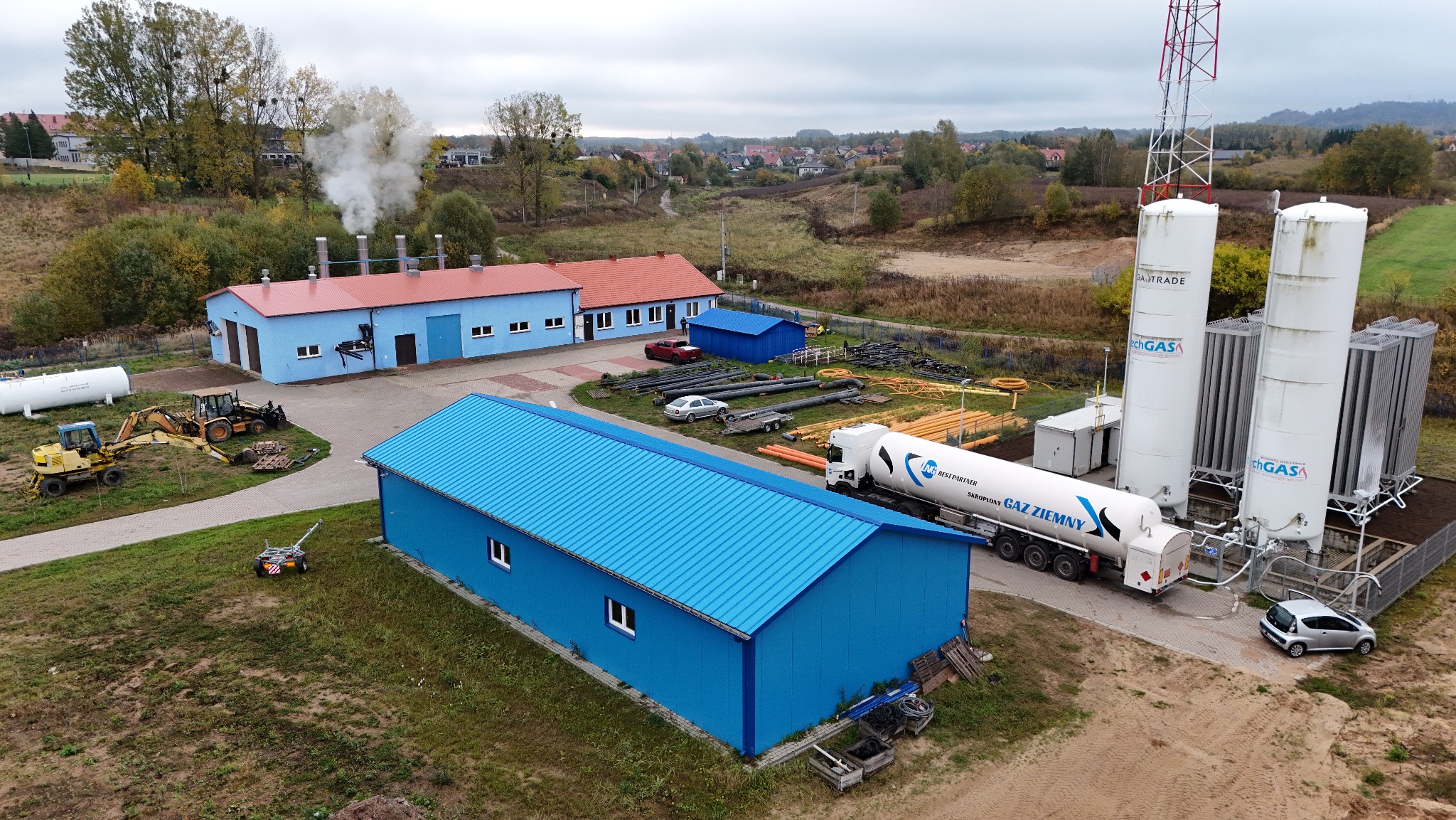Widok z góry na stację gazową z cysterną, zbiornikami i budynkami w niebieskiej kolorystyce. W tle zabudowa wiejska i drzewa.