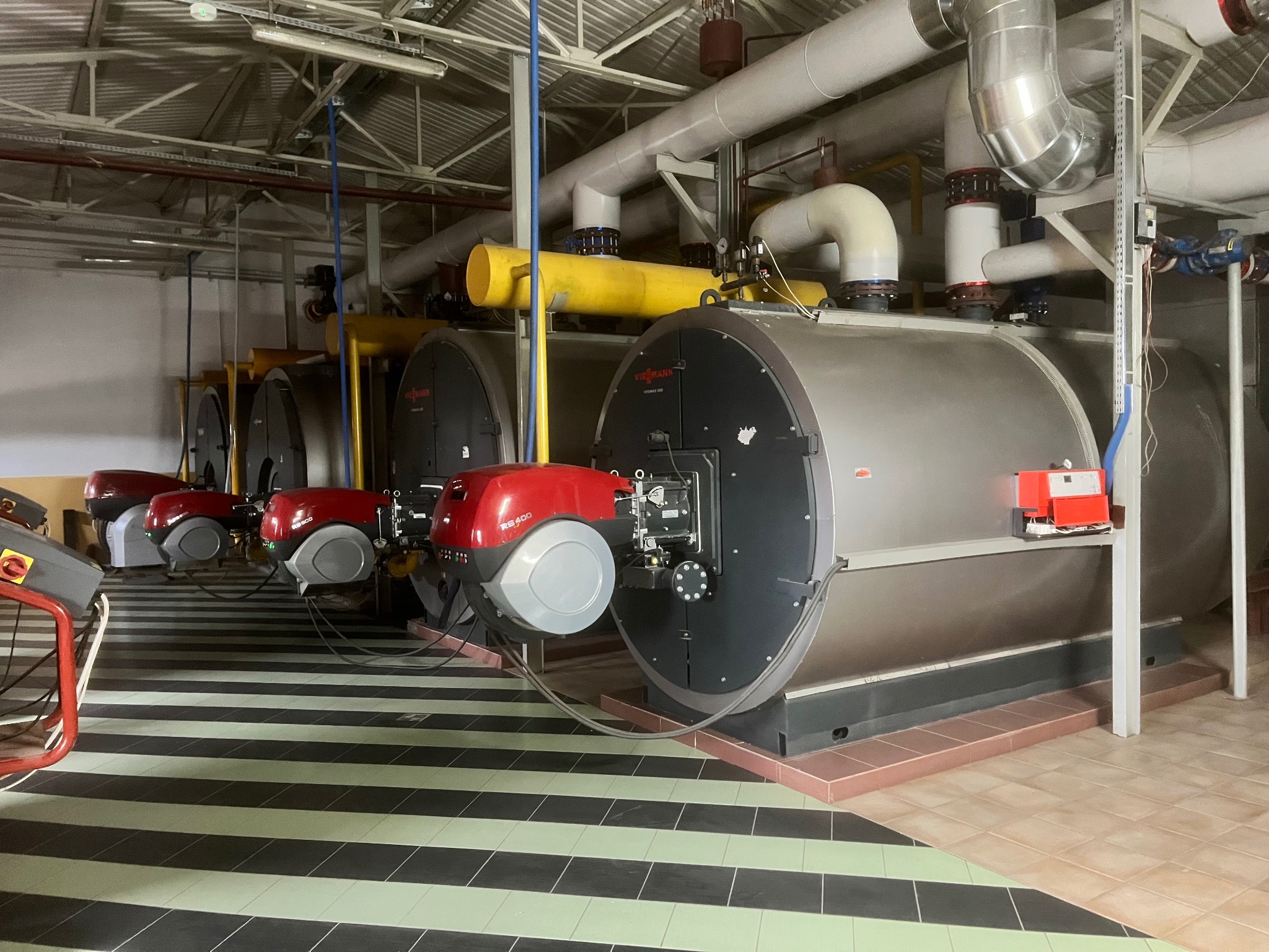 Rząd kotłów gazowych Viessmann z czerwonymi palnikami w kotłowni z rurami i instalacjami pod sufitem. Podłoga w czarno-zielone pasy, ściany białe.