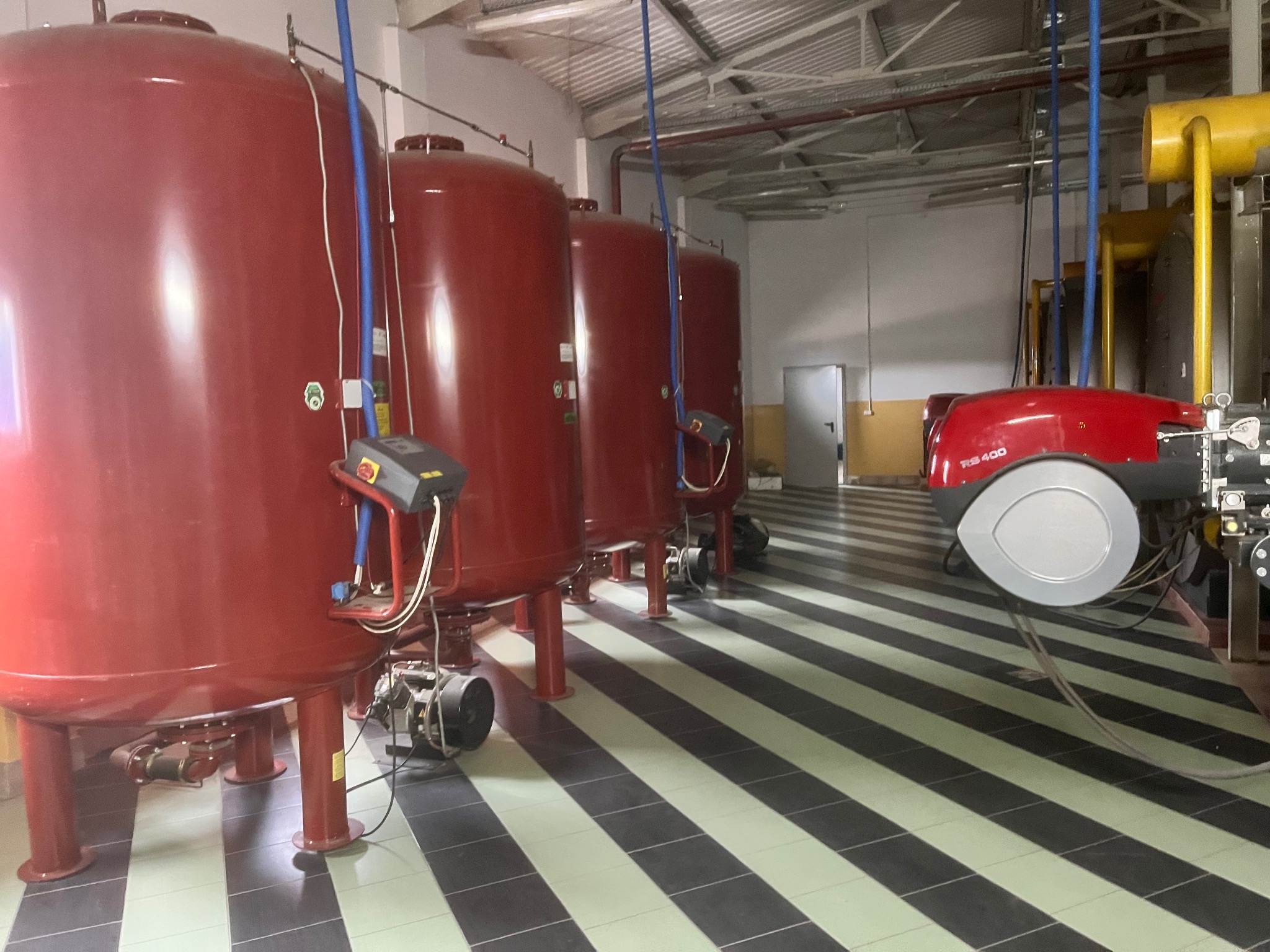Czerwone zbiorniki akumulacyjne ciepła w kotłowni z piecem gazowym RS 400, na podłodze płytki w czarno-białe pasy. Widoczne rury i instalacje.