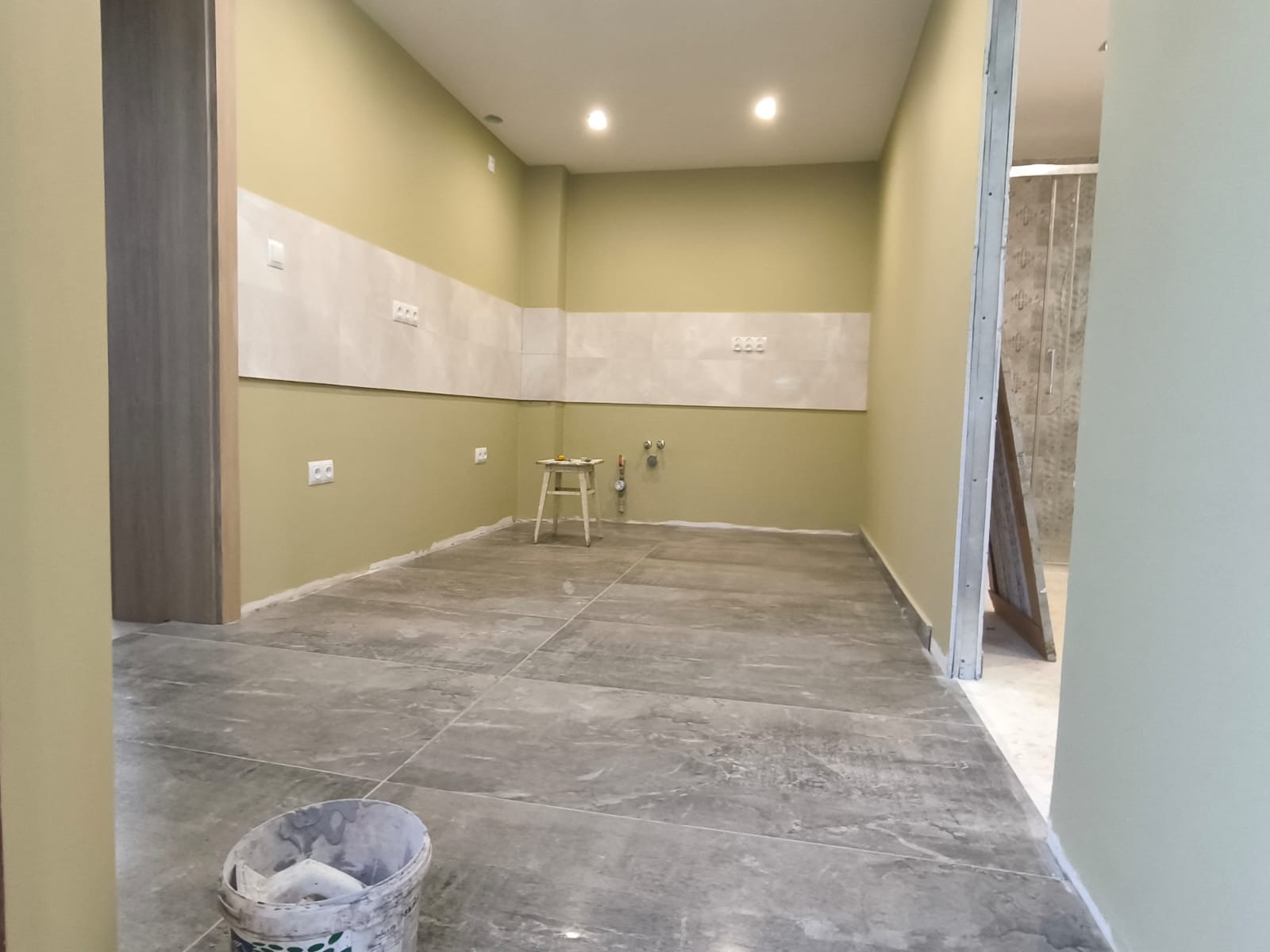 Wnętrze w trakcie wykańczania: szare płytki podłogowe, fragment ściany z białymi płytkami, widoczne gniazdka elektryczne i rury. W tle drzwi do łazienki.