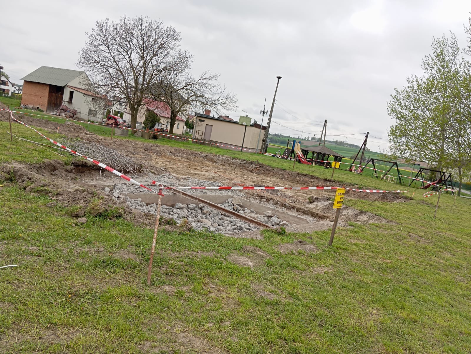 Teren budowy z wykopem fundamentowym, zabezpieczony taśmą ostrzegawczą. W tle budynki mieszkalne i plac zabaw. Widok na plac budowy w pochmurny dzień.