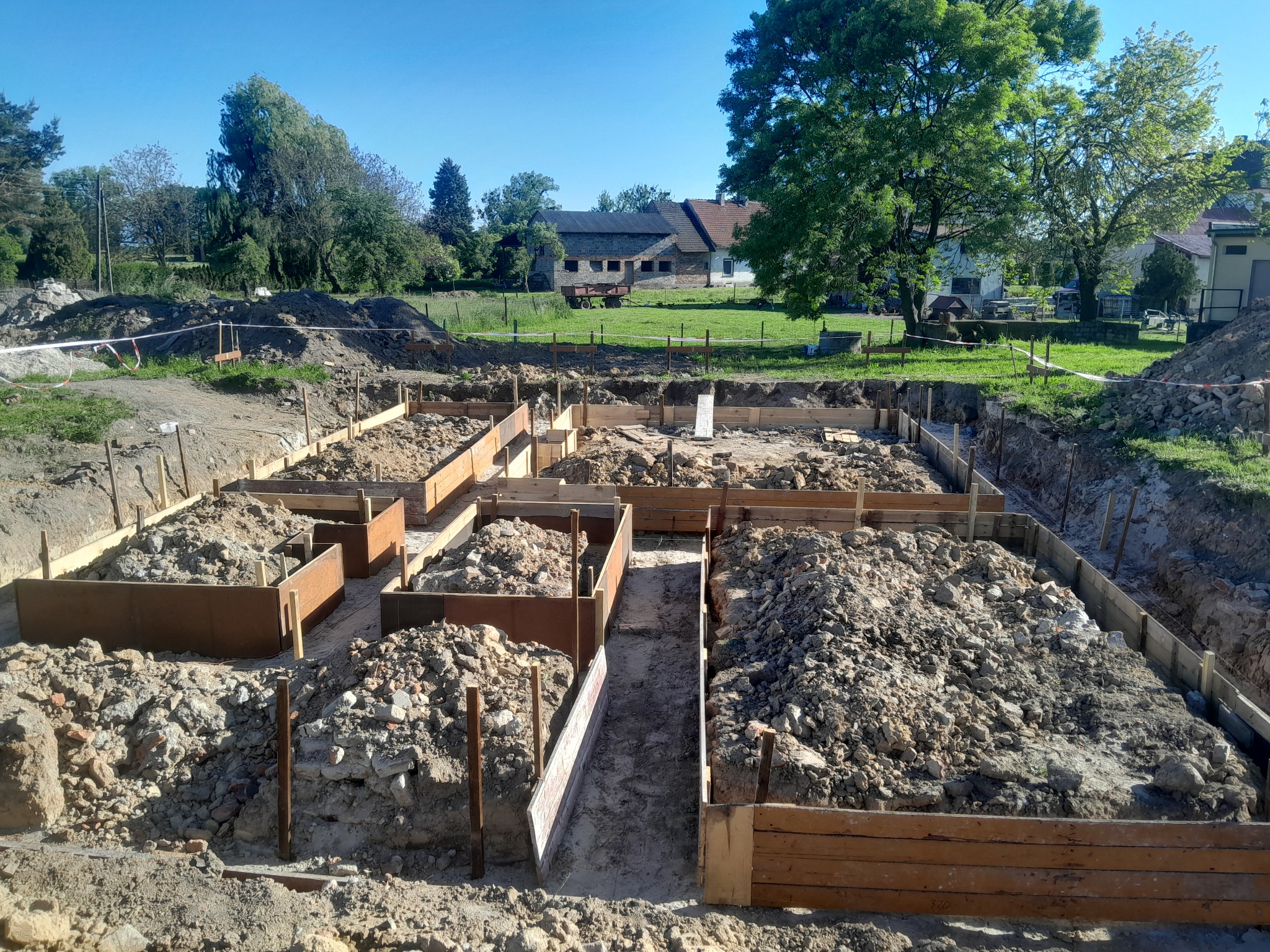 Wykonane deskowanie pod fundamenty domu jednorodzinnego, wypełnione ziemią i gruzem. W tle zielony krajobraz z zabudowaniami. Słoneczny dzień.