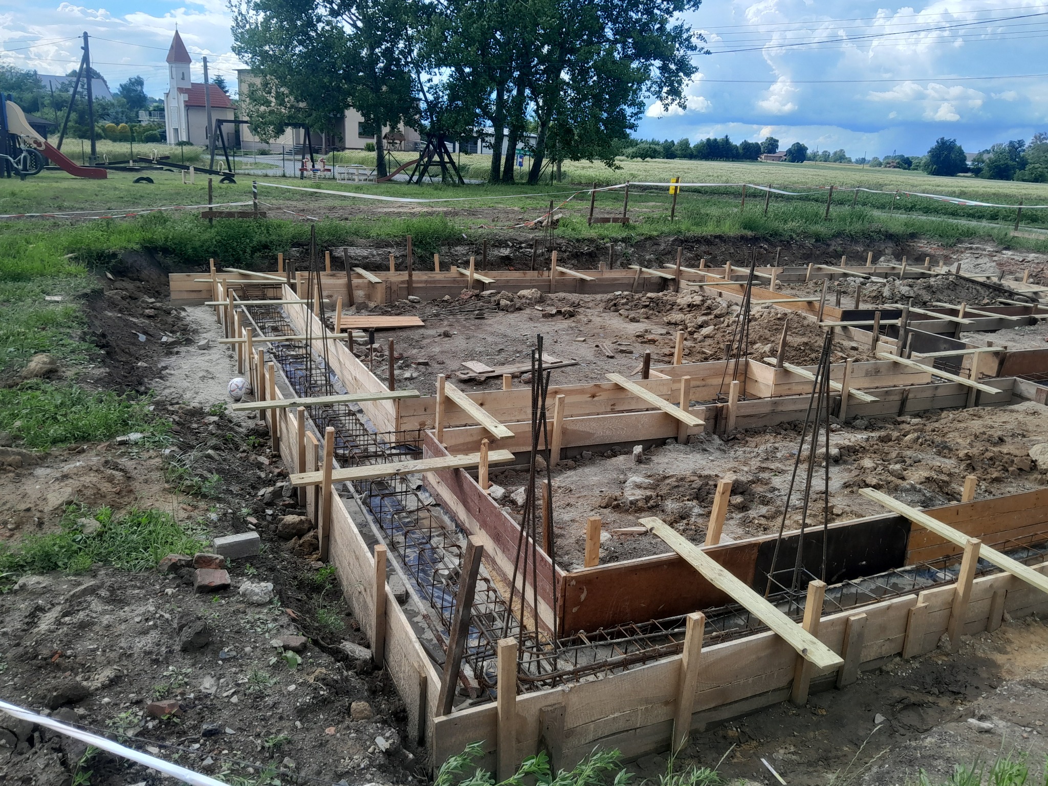 Zbrojenie fundamentów w drewnianym szalunku na placu budowy. W tle widoczny kościół i plac zabaw, co sugeruje lokalizację w pobliżu terenów rekreacyjnych.