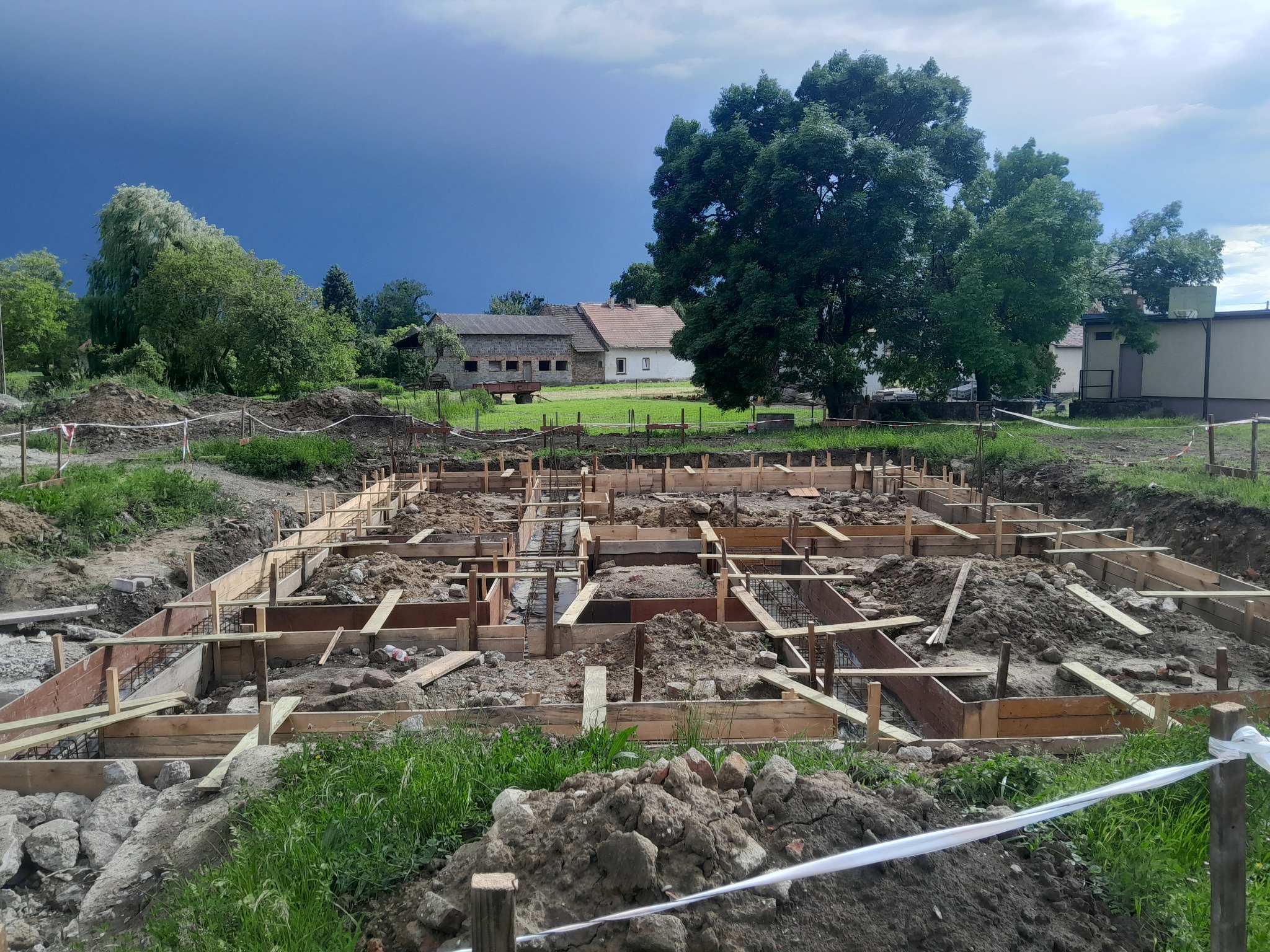 Szeroki kadr na drewniane szalunki fundamentów domu jednorodzinnego w trakcie budowy, z widokiem na wiejski krajobraz i burzowe chmury w tle.