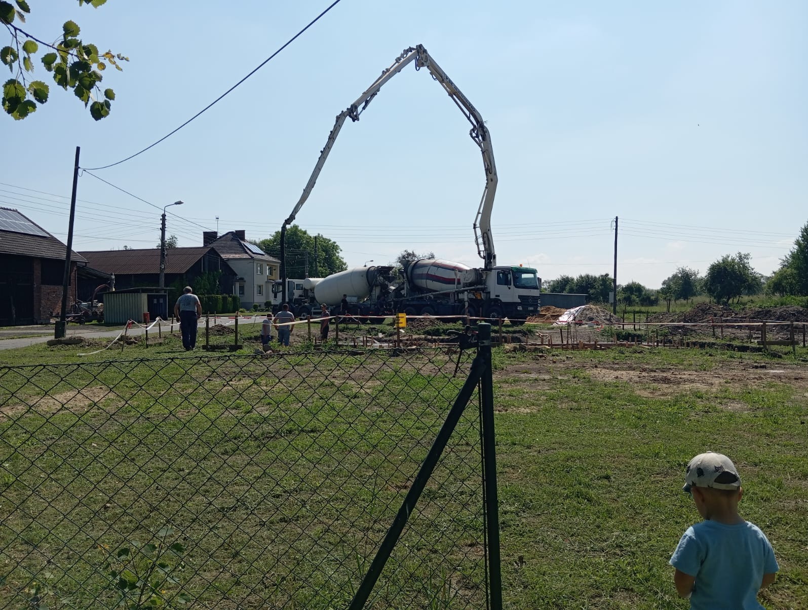 Betoniarka z pompą wylewa beton na fundamenty domu jednorodzinnego. Widoczne szalunki i zbrojenie. Na pierwszym planie siatka ogrodzeniowa i dziecko.