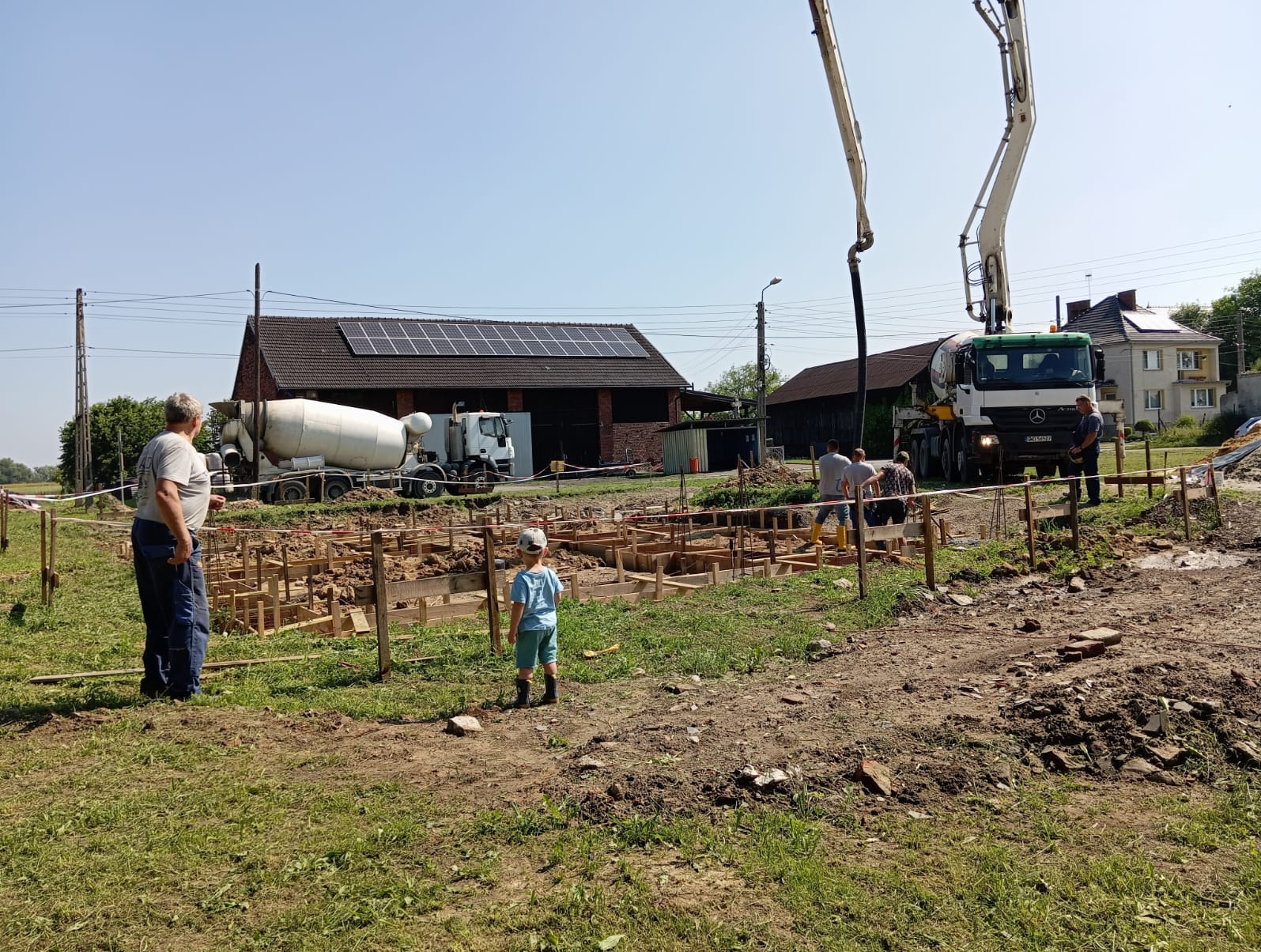 Fundamenty domu w trakcie budowy: drewniane szalunki, ekipa budowlana, pompy do betonu i betoniarki na tle wiejskiego krajobrazu w słoneczny dzień. Dziecko obserwuje prace.