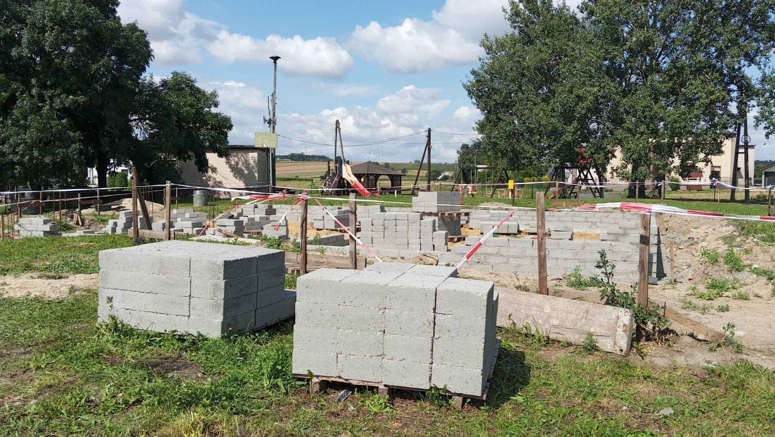 Budowa domu z bloczków betonowych na działce z zieloną trawą, zabezpieczona taśmą ostrzegawczą. Widoczne fundamenty i stos bloczków na palecie.