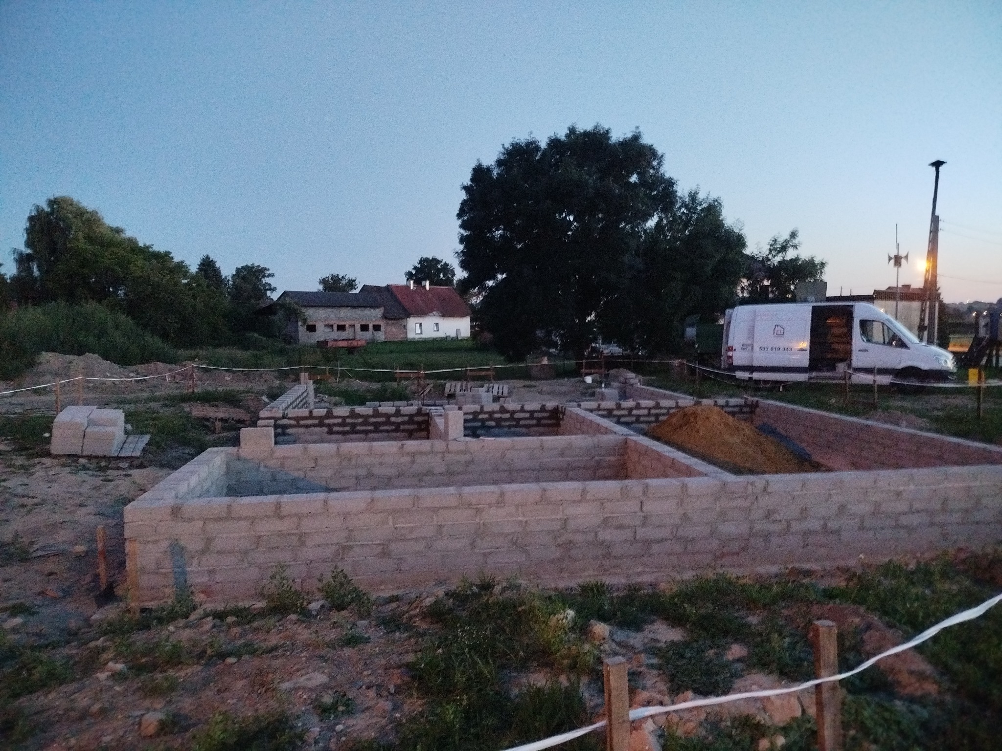 Fundamenty domu z bloczków betonowych w trakcie budowy, widoczny biały bus na działce, w tle zabudowania gospodarcze i drzewa, wieczorny krajobraz.