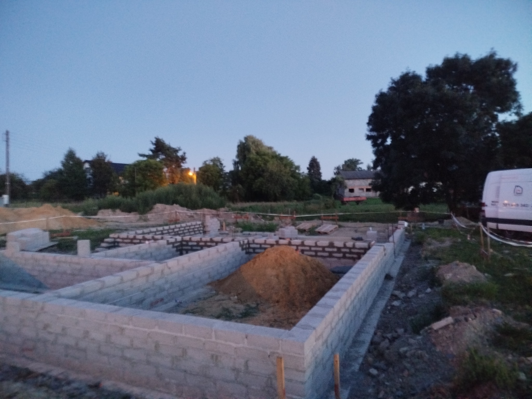 Fundamenty domu z bloczków betonowych w trakcie budowy, widok z góry, wieczorne oświetlenie, w tle zieleń i budynek gospodarczy.