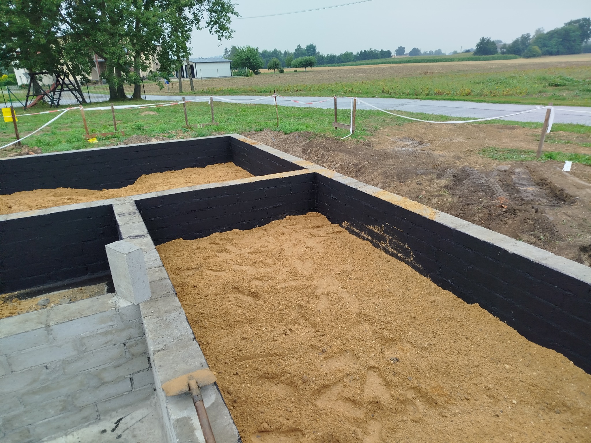 Fundamenty domu w trakcie budowy: ściany z izolacją bitumiczną, wypełnione piaskiem. Widok z góry na plac budowy z zielonym tłem.