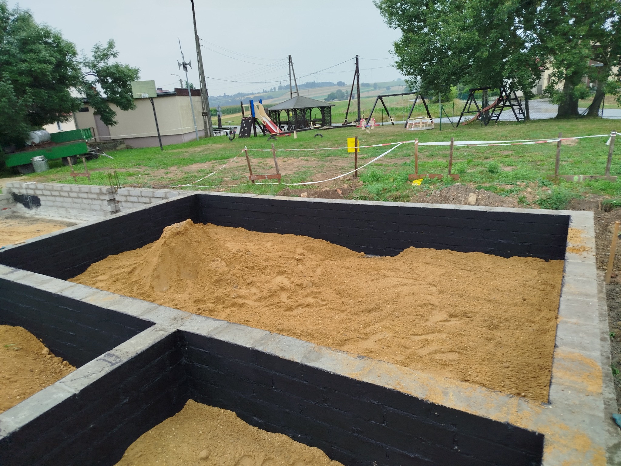 Fundamenty domu w trakcie budowy, zabezpieczone czarną izolacją, wypełnione piaskiem. W tle plac zabaw i zieleń.