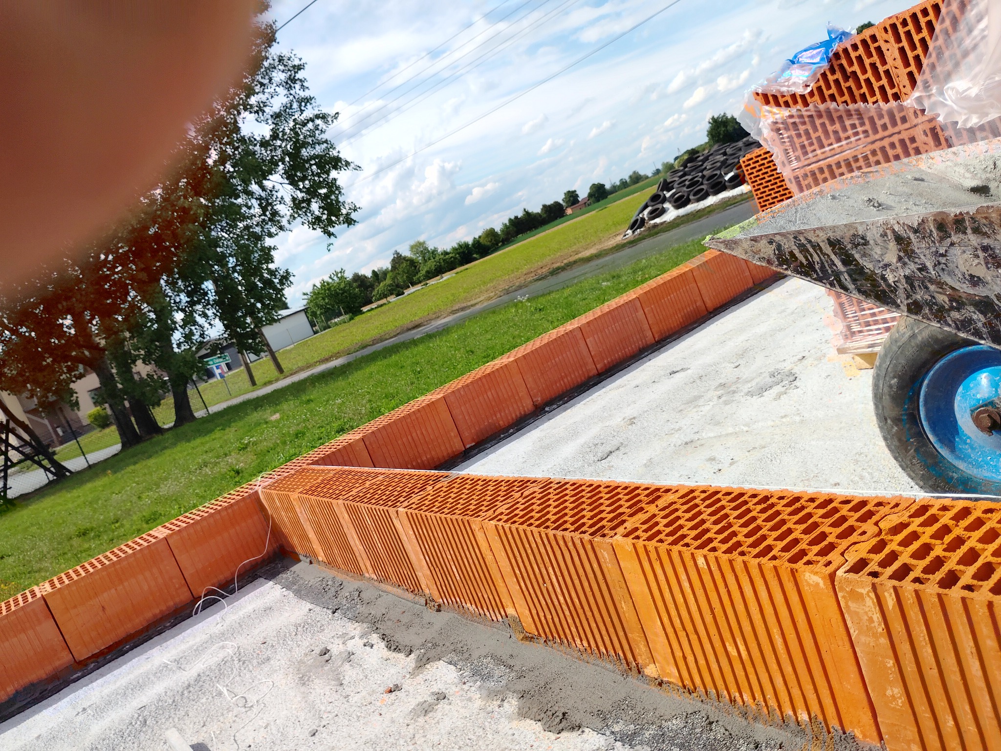 Wznoszenie ścian fundamentowych z bloczków ceramicznych na betonowej płycie. Widoczna taczka z zaprawą murarską i paleta z bloczkami. Trawa i drzewa w tle.