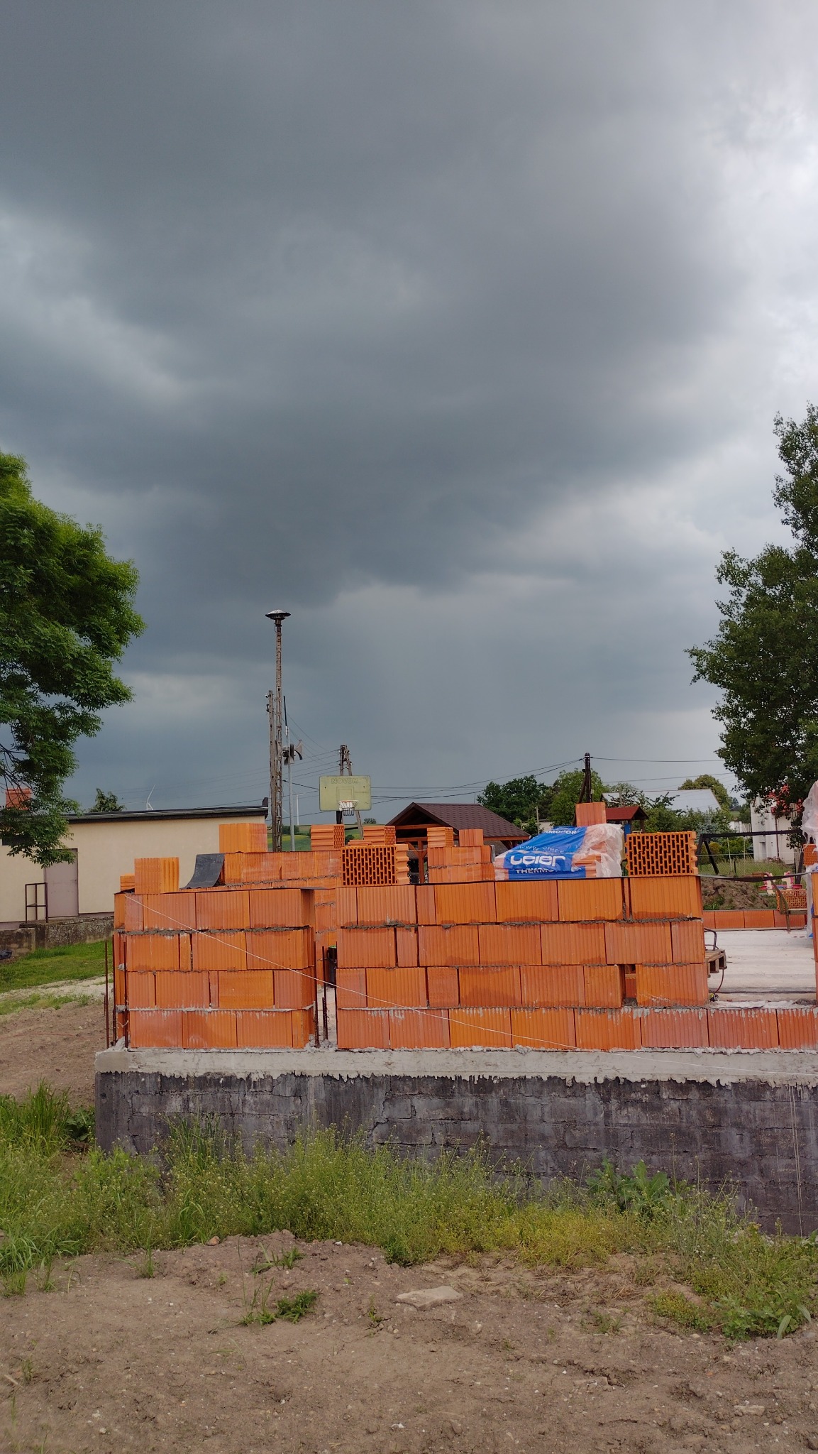 Wznoszone ściany z czerwonej cegły ceramicznej na fundamencie. W tle pochmurne niebo i elementy zabudowy wiejskiej. Częściowo widoczne materiały budowlane.