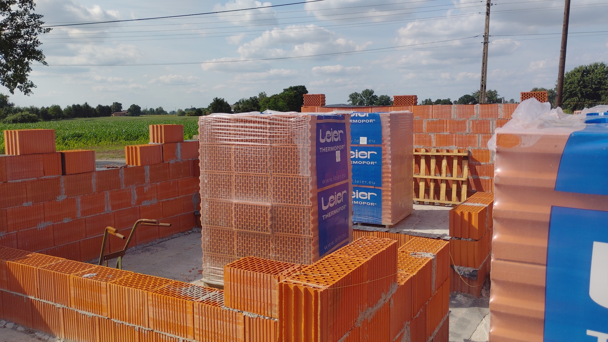 Wznoszone ściany z czerwonej cegły ceramicznej Leier Thermopor, palety z cegłami w folii, widok na budowę domu w słoneczny dzień, zielone pola w tle.
