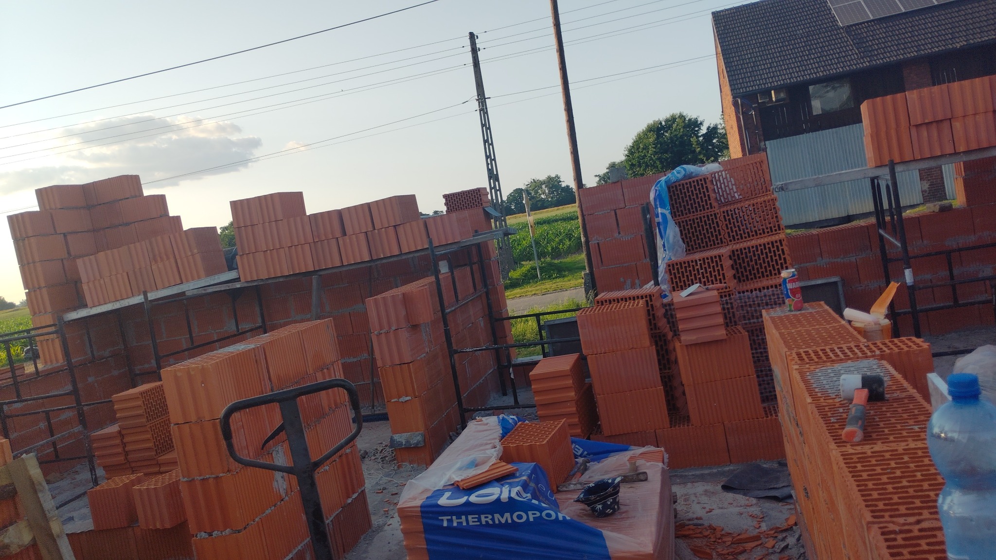 Cegły ceramiczne Leier Thermopor na paletach i rusztowaniach, przygotowane do budowy domu. Widoczne narzędzia murarskie i butelka wody. W tle budynek z panelami słonecznymi.