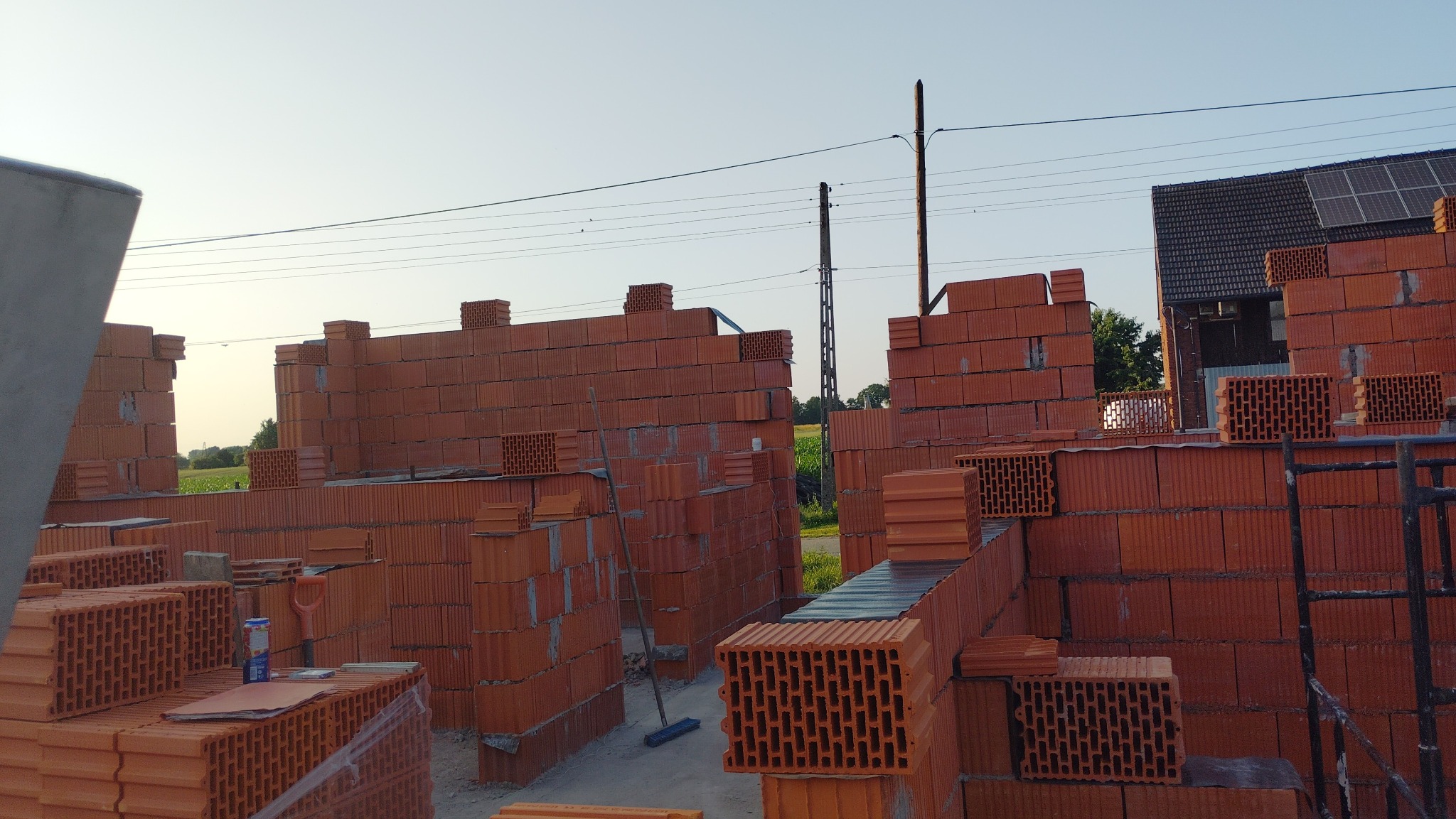 Wznoszenie ścian domu z czerwonej cegły ceramicznej. Widoczne fragmenty ścian, rusztowanie, narzędzia i materiały budowlane. W tle dom z panelami słonecznymi i zielone pole.