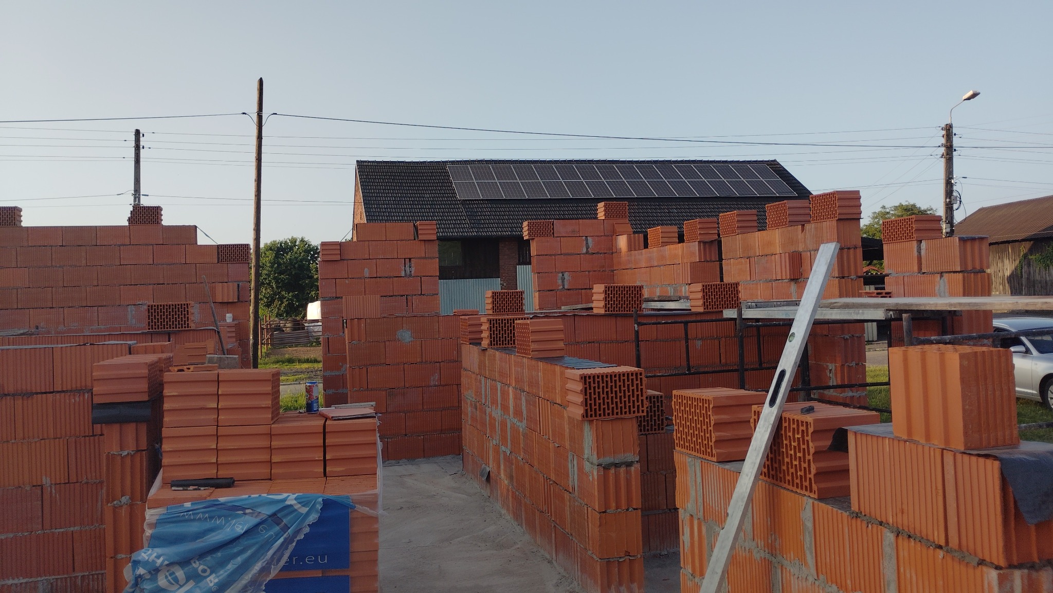 Wznoszone mury z czerwonej cegły ceramicznej na budowie domu jednorodzinnego. Widoczny poziomica, palety z cegłami i dom z panelami słonecznymi w tle.