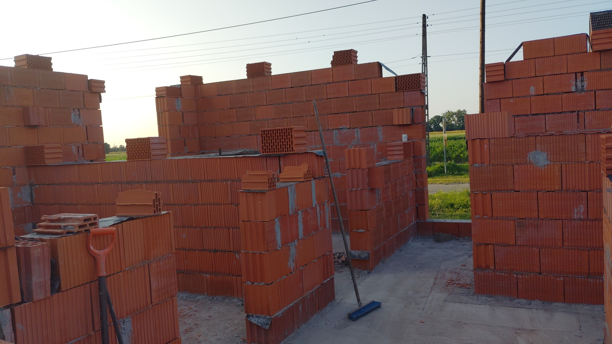 Wznoszenie ścian domu z czerwonej cegły ceramicznej. Widoczne narzędzia murarskie: łopata i zmiotka. Prace murarskie w toku, widok na zewnątrz budynku.