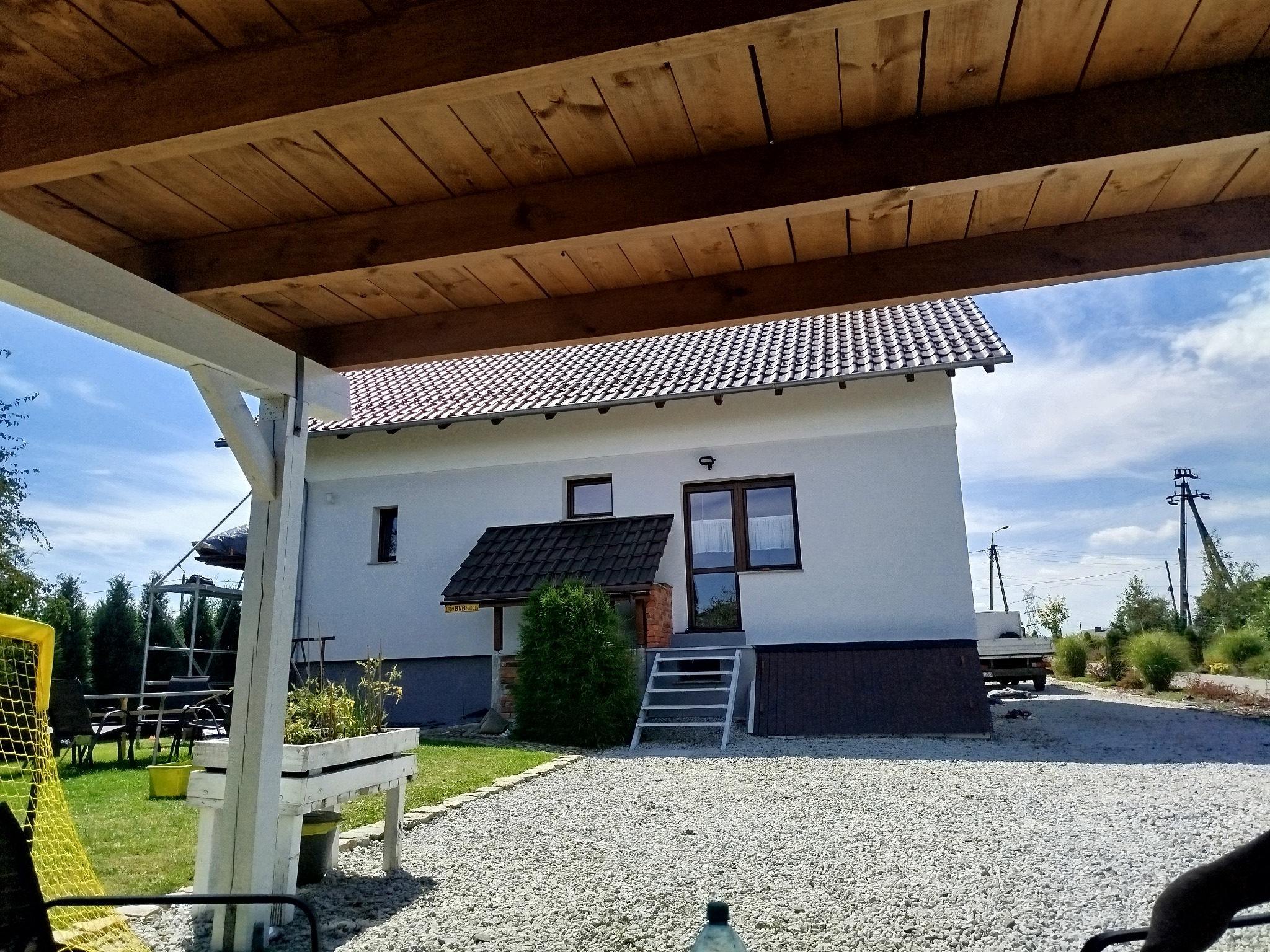 Biały dom z brązowym dachem widziany spod drewnianej konstrukcji z białymi słupami. Podjazd wysypany żwirem, zieleń w tle.