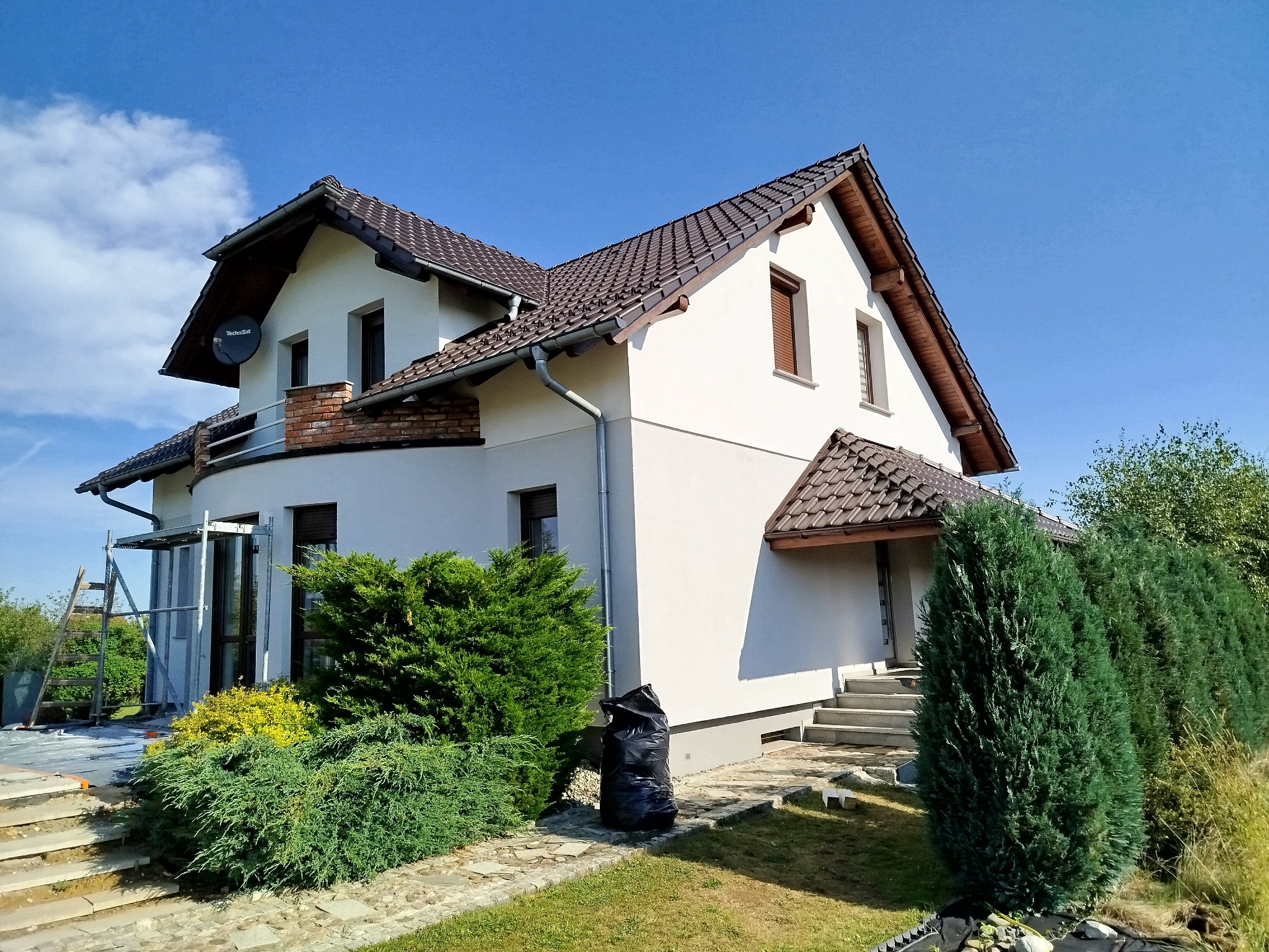 Biały dom jednorodzinny z brązowym dachem w słoneczny dzień. Widoczne rusztowania i worki na śmieci sugerują prace wykończeniowe.