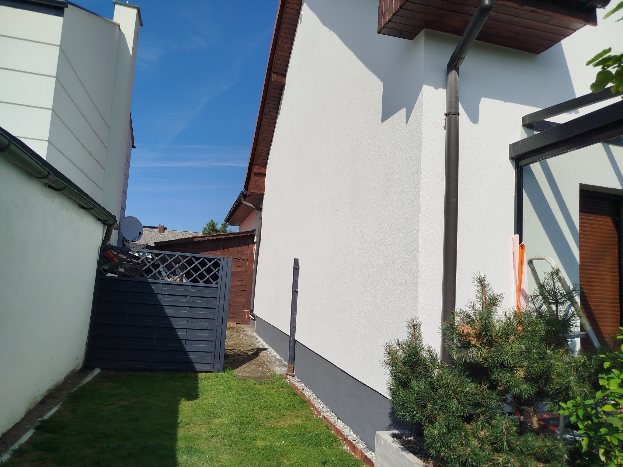 Boczny widok otynkowanego domu z szarą podmurówką, rynną i fragmentem ogrodu. Widoczny fragment ogrodzenia i sąsiedni budynek w tle, błękitne niebo.