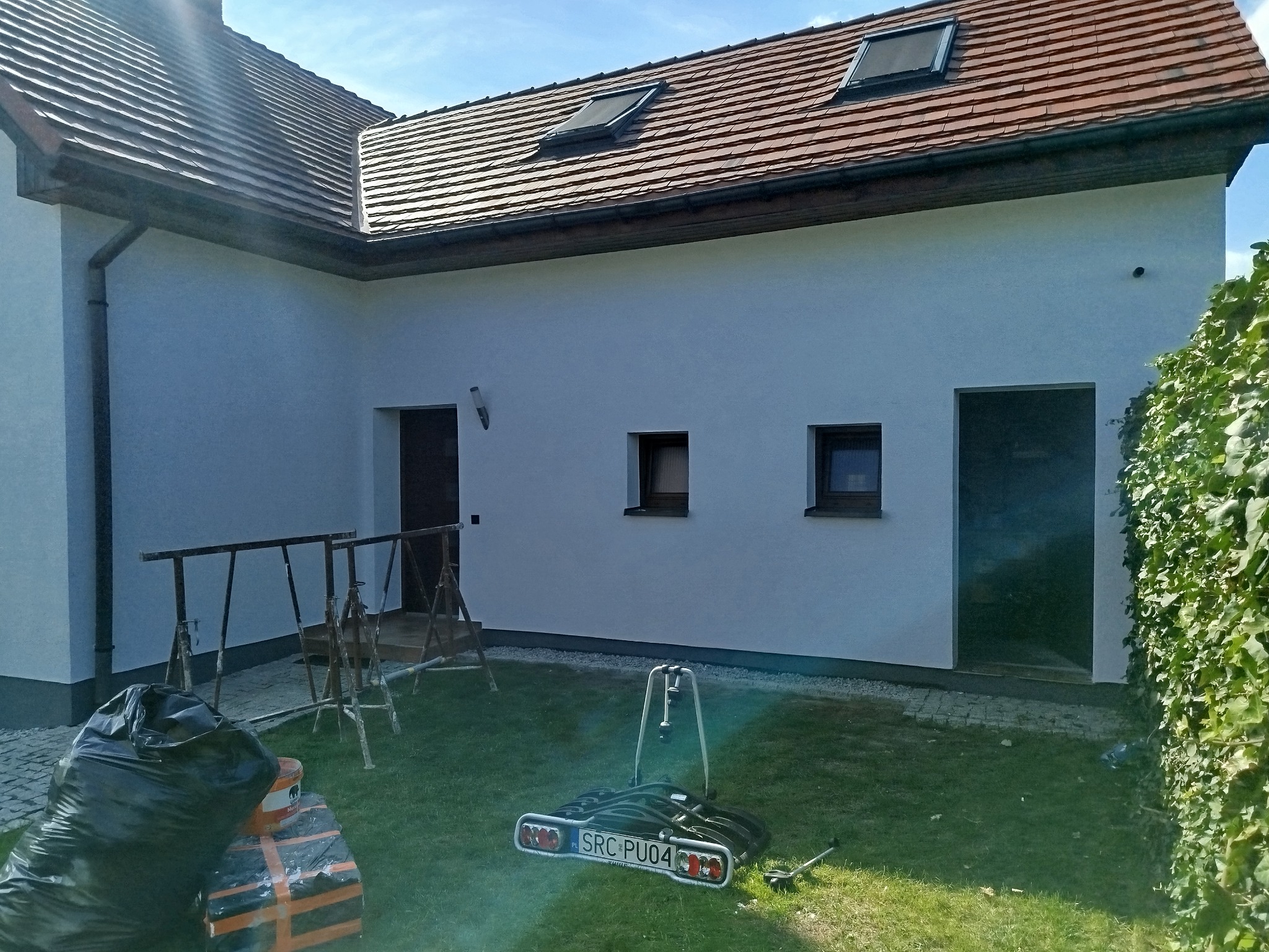 Biała elewacja domu z czerwonym dachem, widoczne rusztowania i akcesoria malarskie na trawniku. Świeżo pomalowana fasada z oknami i drzwiami.