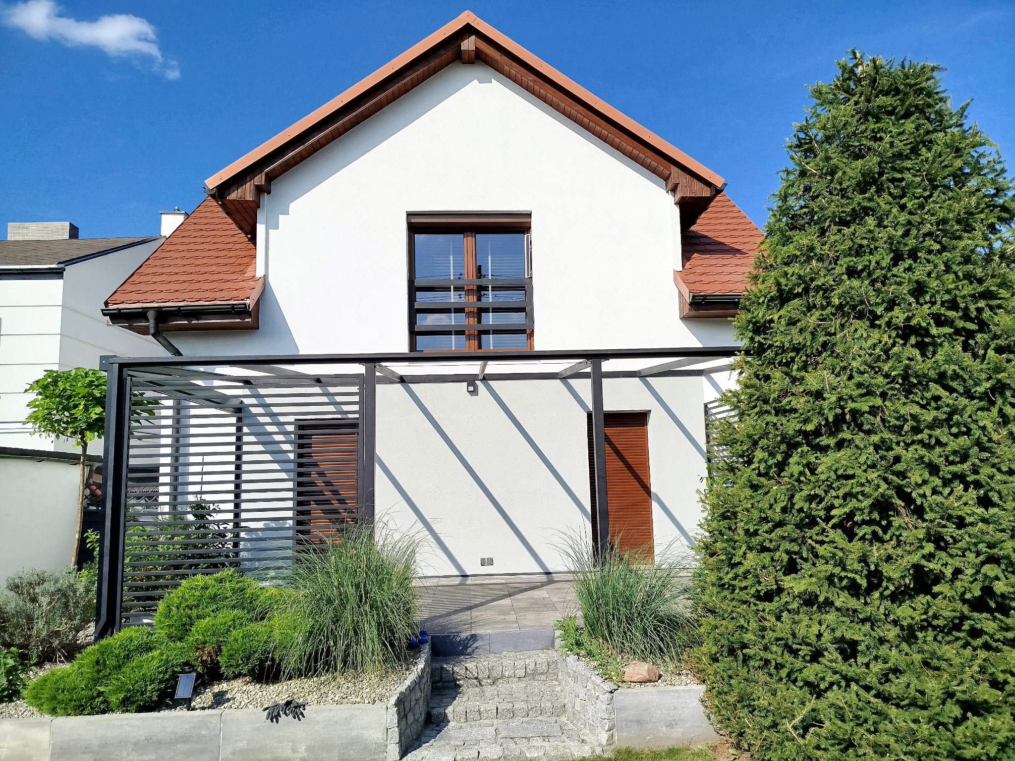 Biały dom z brązowym dachem i pergolą, otoczony zielenią. Widoczne schody wejściowe i starannie zagospodarowany ogród. Słoneczny dzień, błękitne niebo.
