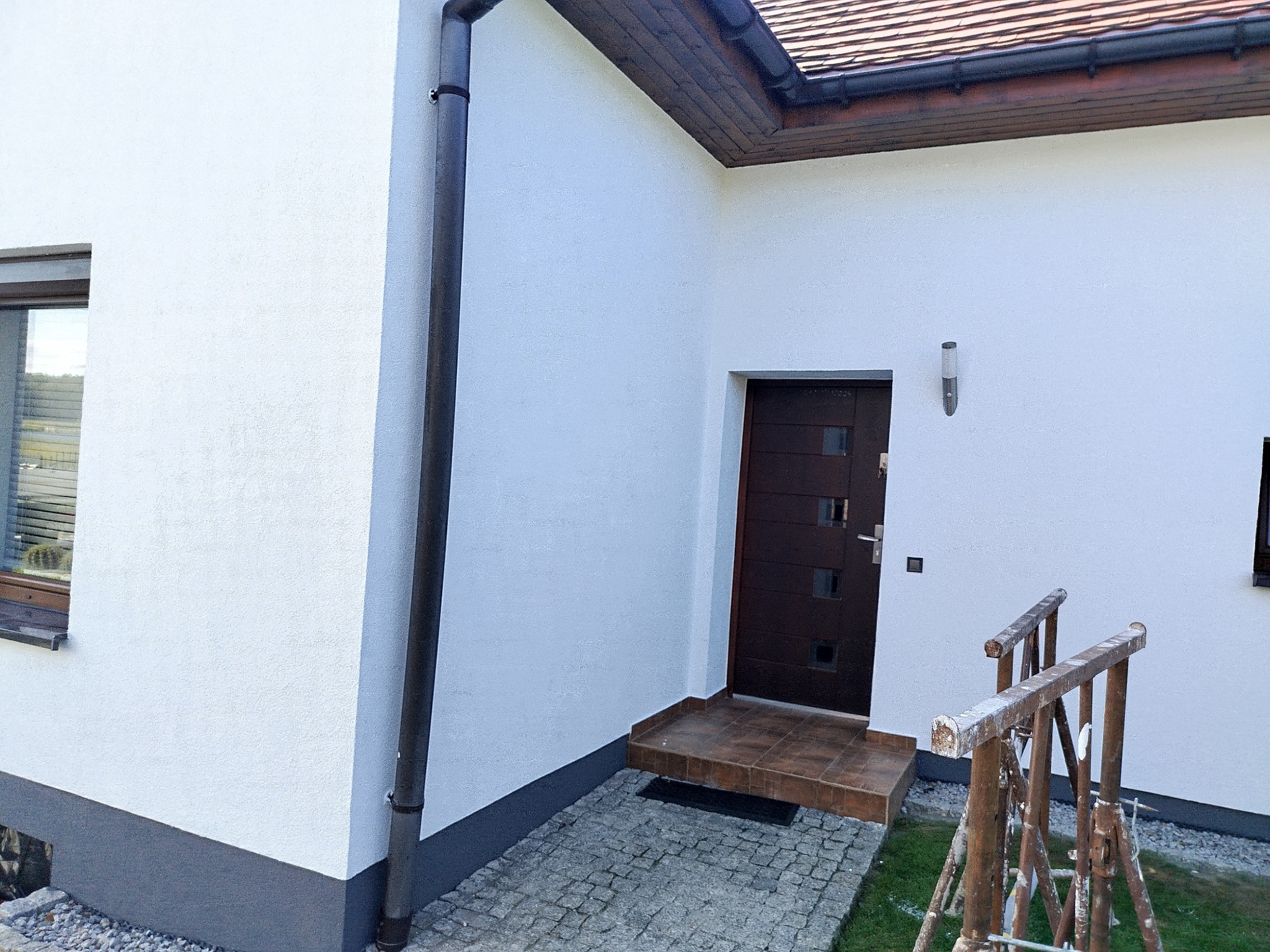 Elewacja domu jednorodzinnego po remoncie: biała ściana z brązowymi akcentami, widoczna rynna, okno i drzwi wejściowe. Na pierwszym planie rusztowanie malarskie.