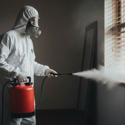 GALPA PAWEŁ BIAŁOŃ - Osoba w kombinezonie ochronnym i masce rozpyla środek dezynfekujący w pomieszczeniu z oknem i żaluzjami. Czerwony zbiornik i dysza w dłoniach.