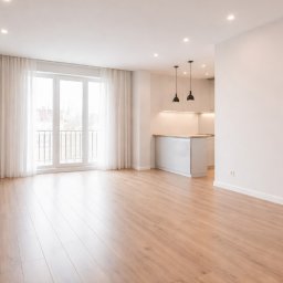 Bud-Max.Air Yurii Tepliuk - Jasne, przestronne wnętrze z drewnianą podłogą, białymi ścianami i widokiem na kuchnię z wyspą. Duże okno balkonowe z białymi zasłonami wpuszcza dużo światła.