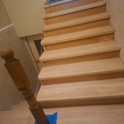 Bud-Max.Air Yurii Tepliuk - Drewniane schody z balustradą zabezpieczoną niebieską taśmą malarską. Widok z góry na jasne stopnie i ciemniejszy element balustrady. W tle widoczne drzwi i okno.