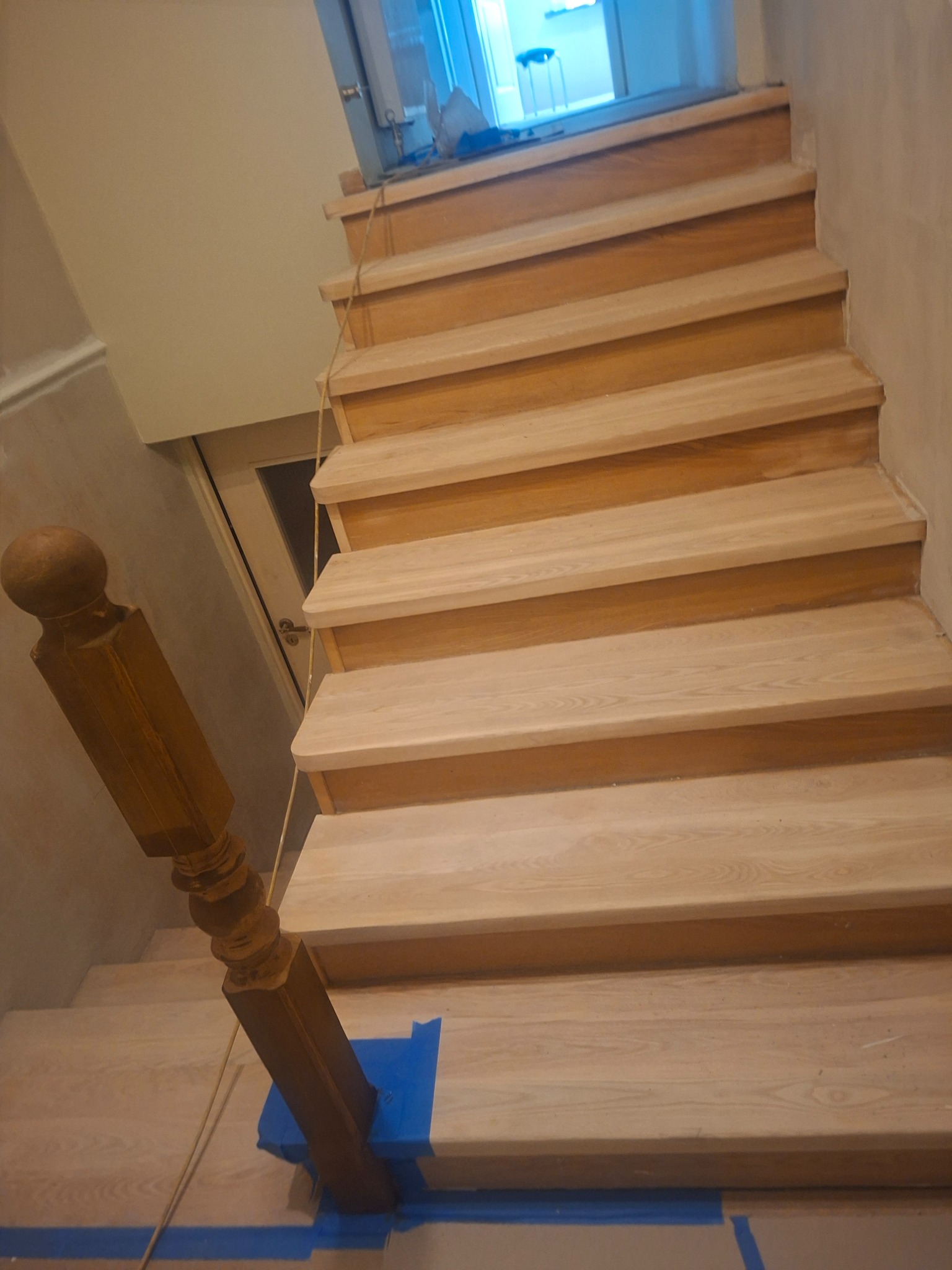 Drewniane schody z balustradą zabezpieczoną niebieską taśmą malarską. Widok z góry na jasne stopnie i ciemniejszy element balustrady. W tle widoczne drzwi i okno.