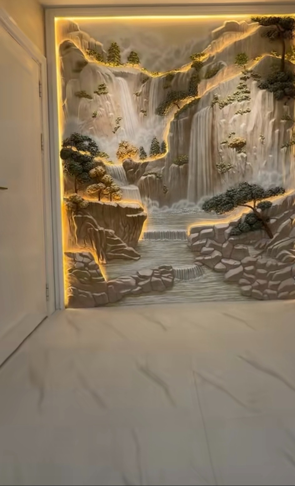 Relief ścienny z motywem wodospadu, podświetlony ciepłym światłem LED, tworzący trójwymiarowy efekt. Dekoracja w jasnych, naturalnych barwach.