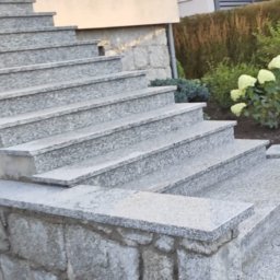 Kamieniarstwo Artystyczne Jacek Kociołek - Zewnętrzne schody z jasnoszarego granitu, prowadzące do domu. Widoczne detale wykończenia i otoczenie z zielenią. Ujęcie z dołu, podkreślające solidność konstrukcji.