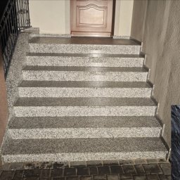 Kamieniarstwo Artystyczne Jacek Kociołek - Zewnętrzne schody z szarego kamienia z balustradą prowadzące do drzwi wejściowych. Widoczny fragment logo firmy kamieniarskiej w prawym dolnym rogu.