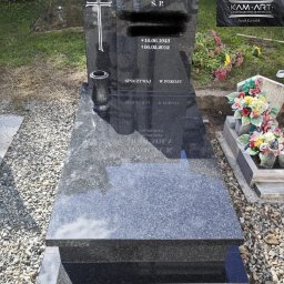 Kamieniarstwo Artystyczne Jacek Kociołek - Czarny, polerowany nagrobek z krzyżem i wazonem, otoczony drobnym żwirem. Widoczne napisy i daty. W tle zieleń i fragment innego nagrobka z kwiatami.