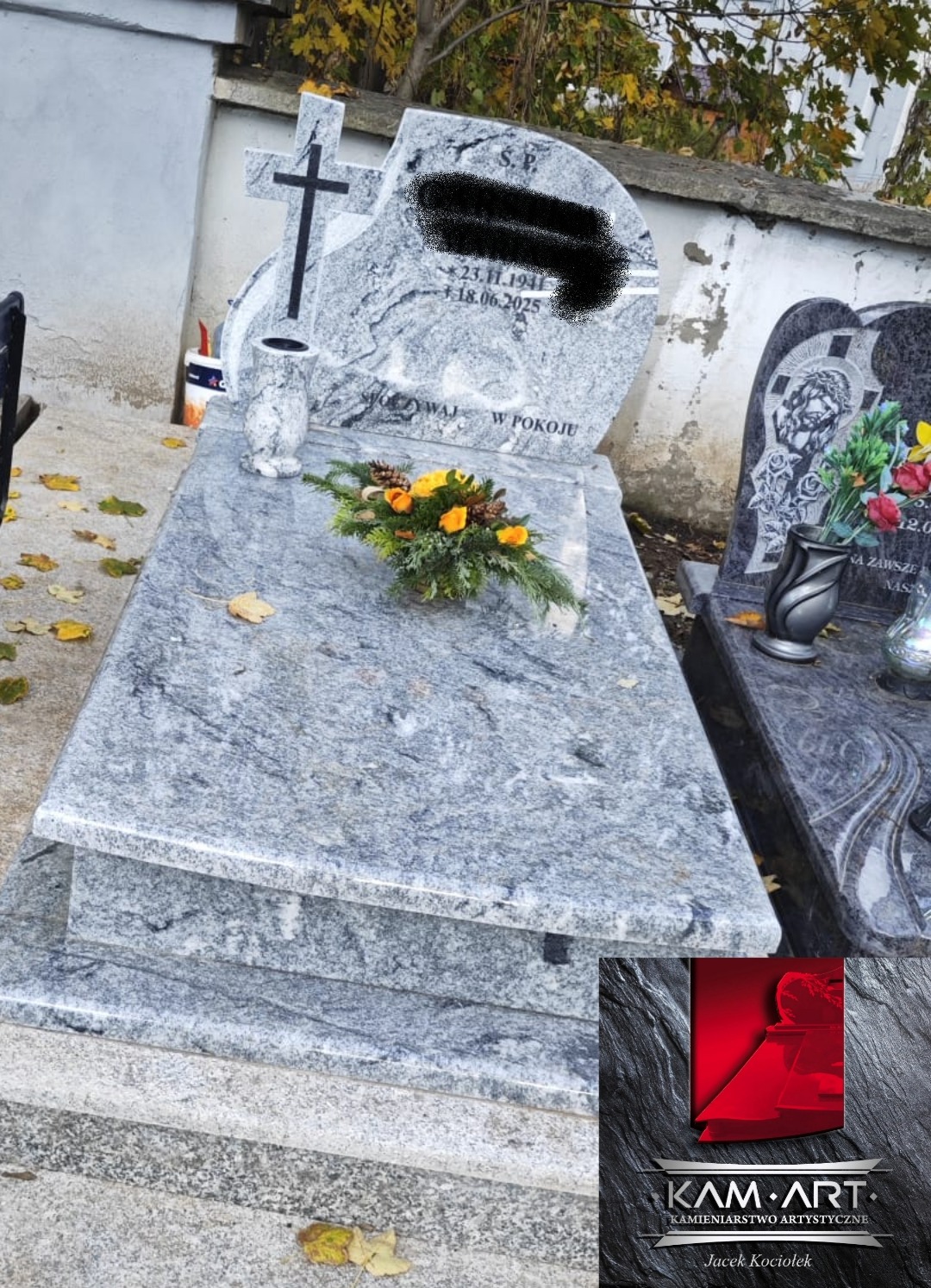 Jasnoszary nagrobek z krzyżem i kwiatami, wykonany z kamienia o wyraźnej fakturze. Widoczny napis 'Spoczywaj w pokoju'. W tle inne nagrobki i jesienne liście.