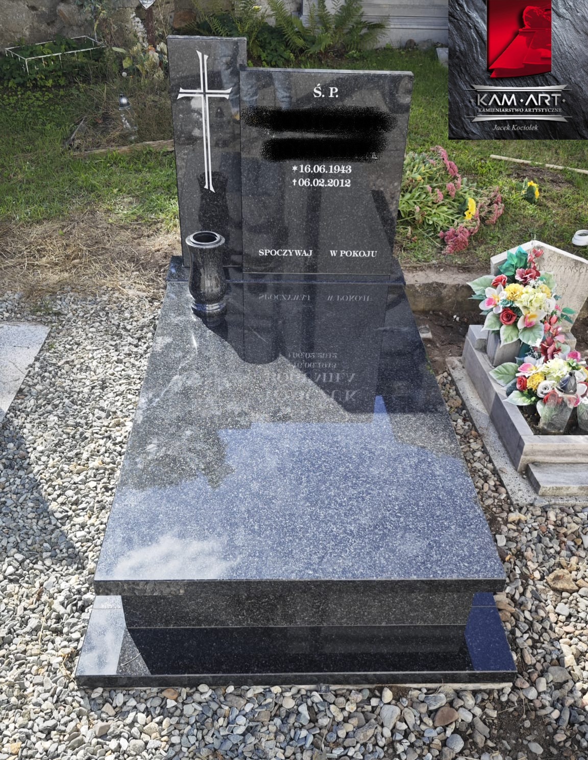 Czarny, polerowany nagrobek z krzyżem i wazonem, otoczony drobnym żwirem. Widoczne napisy i daty. W tle zieleń i fragment innego nagrobka z kwiatami.