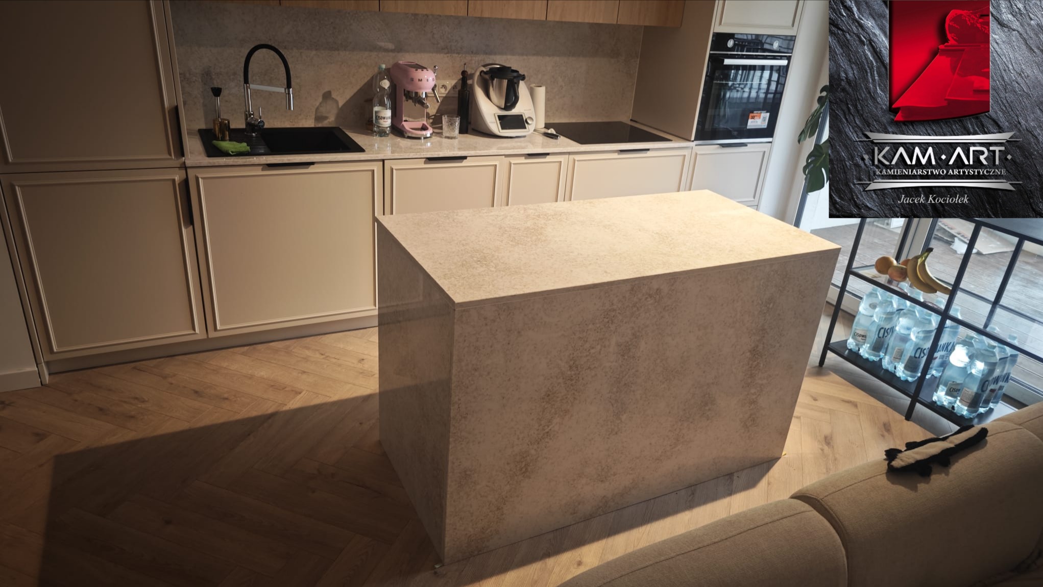 Kamienny blok kuchenny w nowoczesnej kuchni. Beżowy blat z kamienia, drewniana podłoga w jodełkę, minimalistyczny design. W tle logo firmy kamieniarskiej.