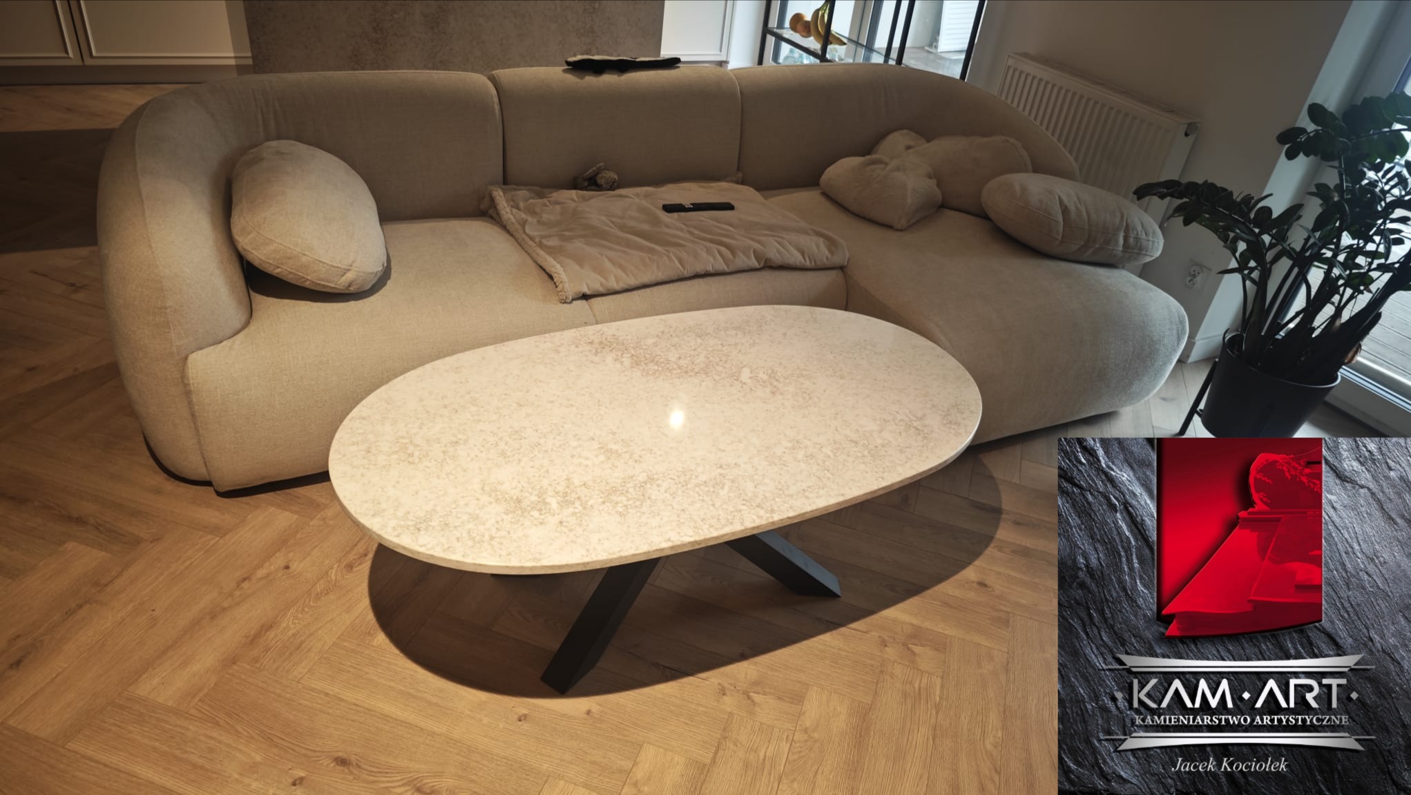 Owalny blat kamienny na czarnych, skośnych nogach, ustawiony w salonie obok beżowej sofy. Widoczny parkiet i logo firmy kamieniarskiej w prawym dolnym rogu.