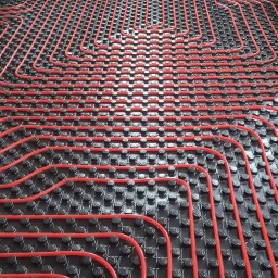 Stanisław Pabiasz INSTAL-DOM - Czerwone rury ogrzewania podłogowego ułożone na czarnej macie izolacyjnej w surowym wnętrzu. Widok z góry na precyzyjny układ pętli grzewczych przed wylewką.