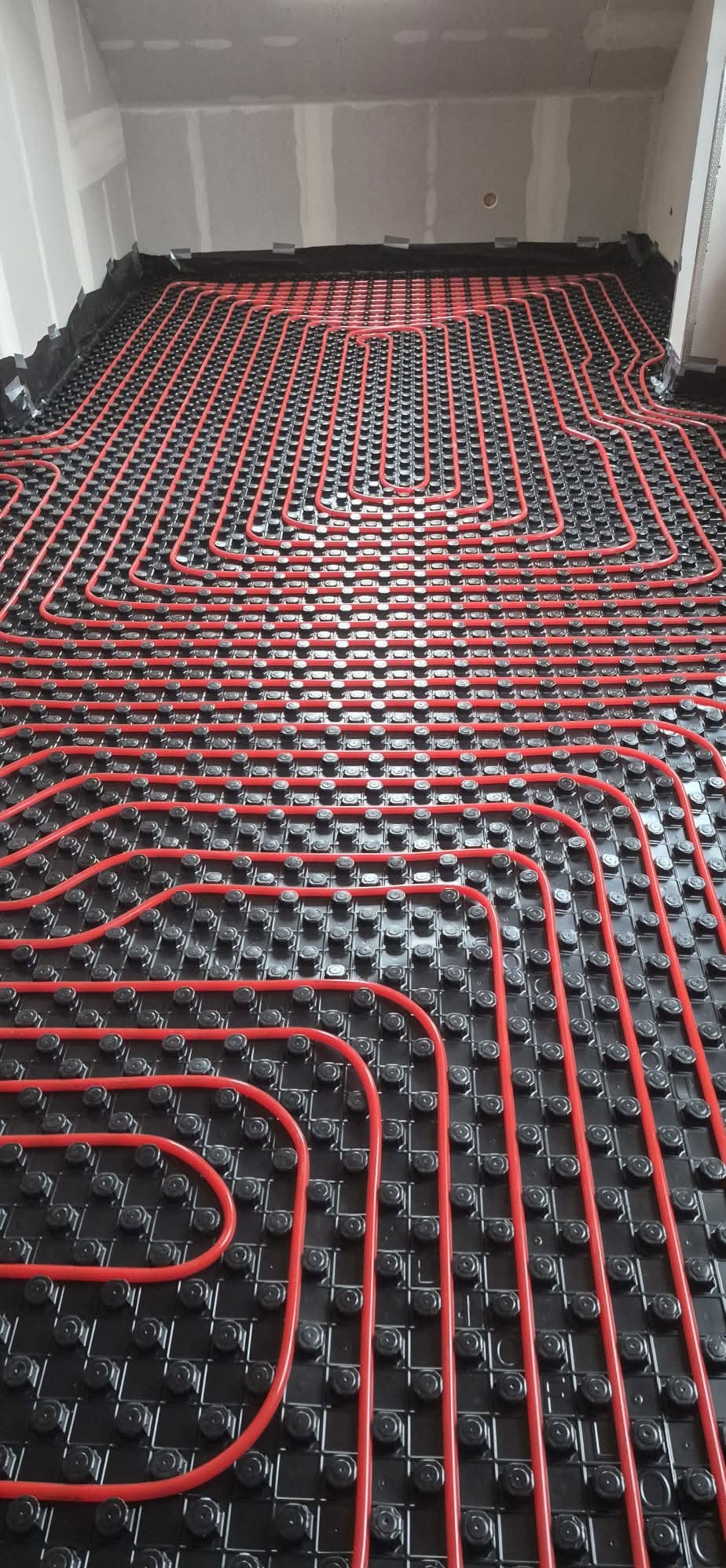 Czerwone rury ogrzewania podłogowego ułożone na czarnej macie izolacyjnej w surowym wnętrzu. Widok z góry na precyzyjny układ pętli grzewczych przed wylewką.