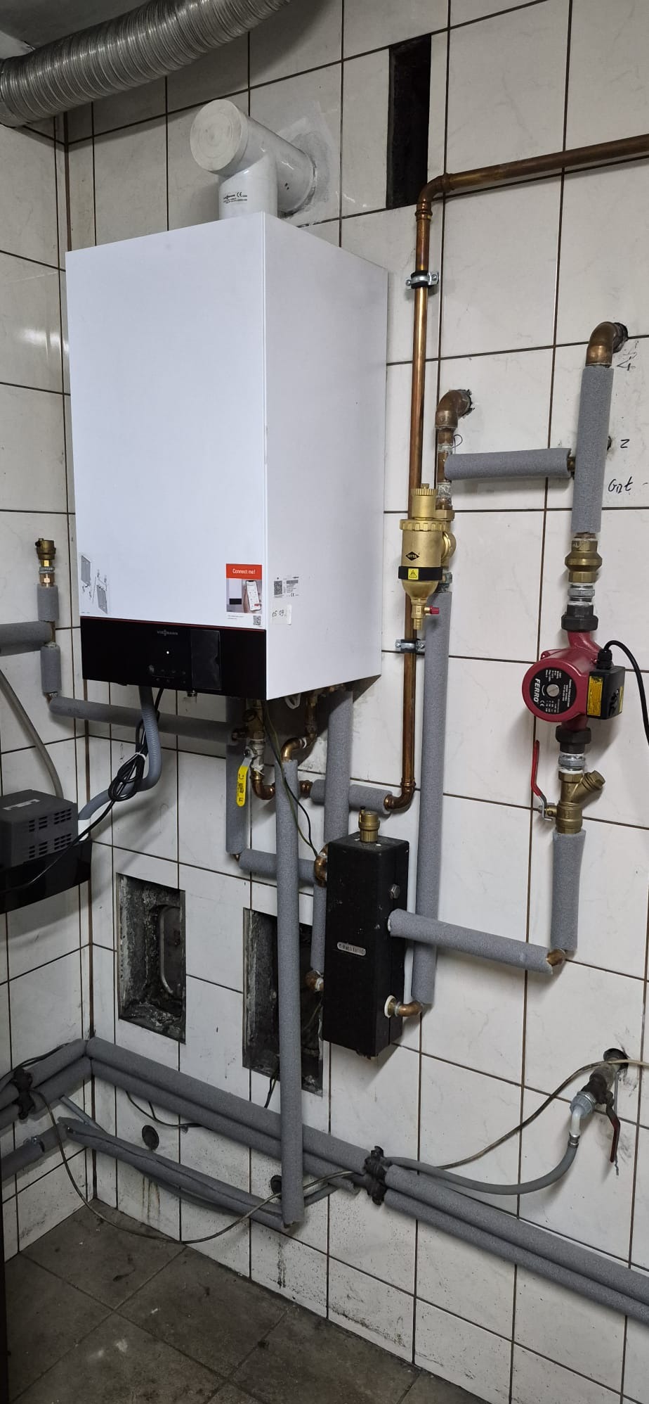 Nowoczesny, biały piec kondensacyjny Viessmann na tle białych płytek. Widoczne rury miedziane i szare izolacje. Elementy instalacji grzewczej w kotłowni po modernizacji.