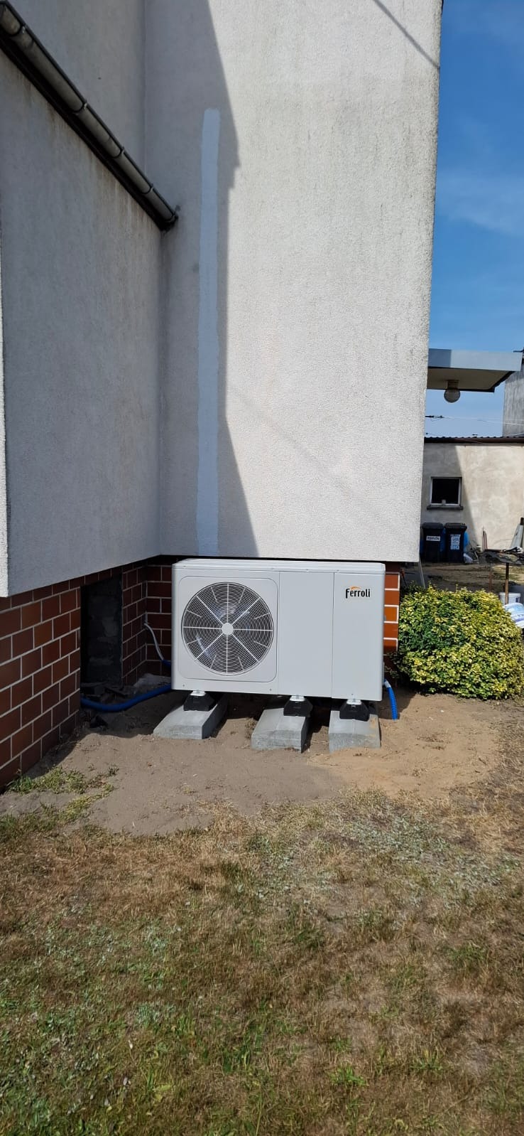 Biała, zewnętrzna pompa ciepła Ferroli zamontowana na betonowych podstawach przy ścianie budynku z ceglaną podmurówką. Widoczne niebieskie rurki. Słoneczny dzień.